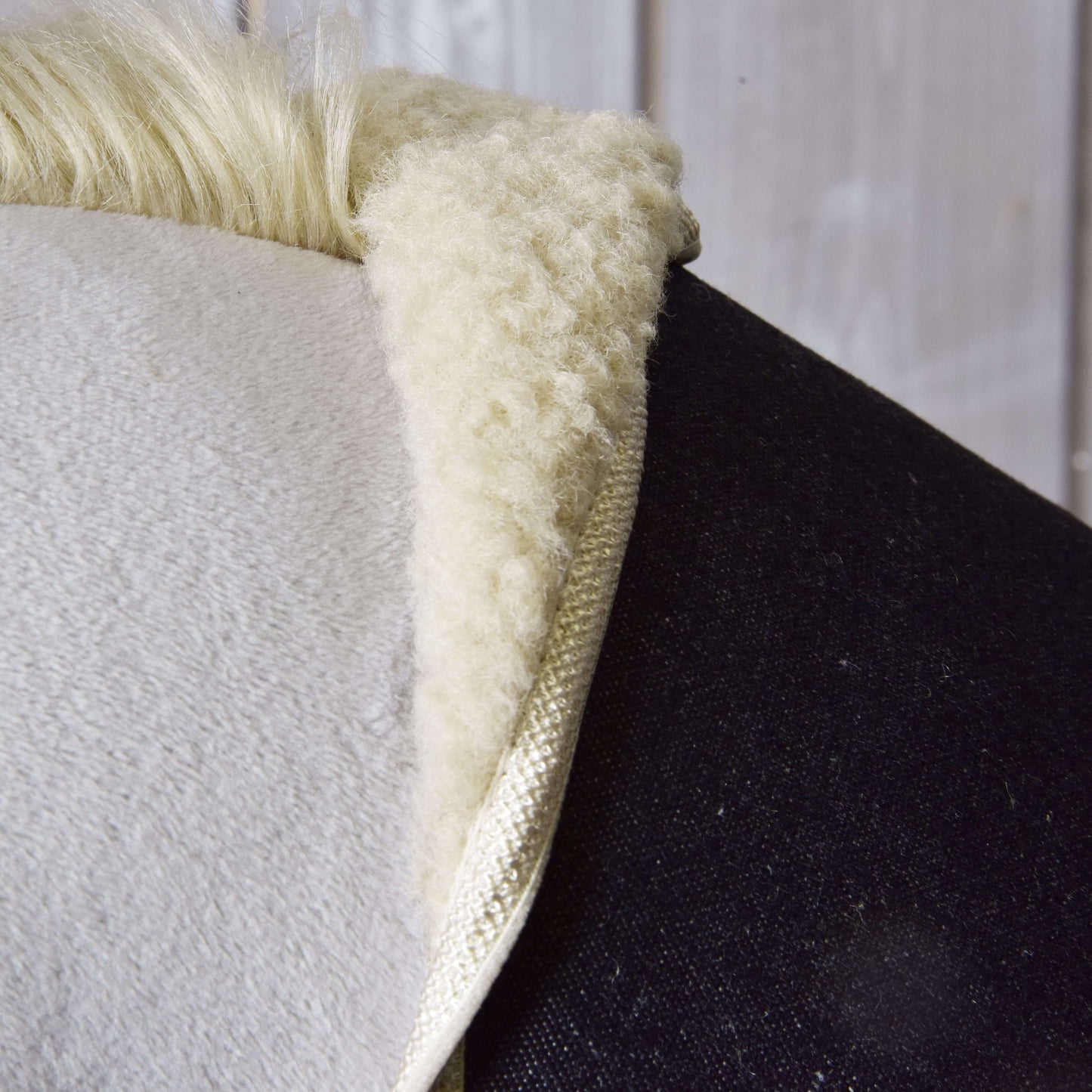 Detailný záber na podsedlovú dečku „Set Blue Denim“ od Hobby Horse LarDen s mäkkou, huňatou bielou textúrou imitujúcou ovčiu kožušinu na chrbte tmavého koňa, s drevenou stenou v pozadí.