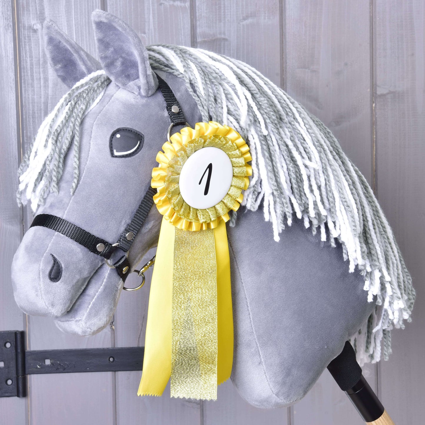 Set Hobby Horse Kokarda NEW od Hobby Horse LarDen so žltou kokardou za prvé miesto označenou „1“ na sivom Hobby Horse s bielou priadzovou hrivou, zobrazený na pozadí drevenej steny.