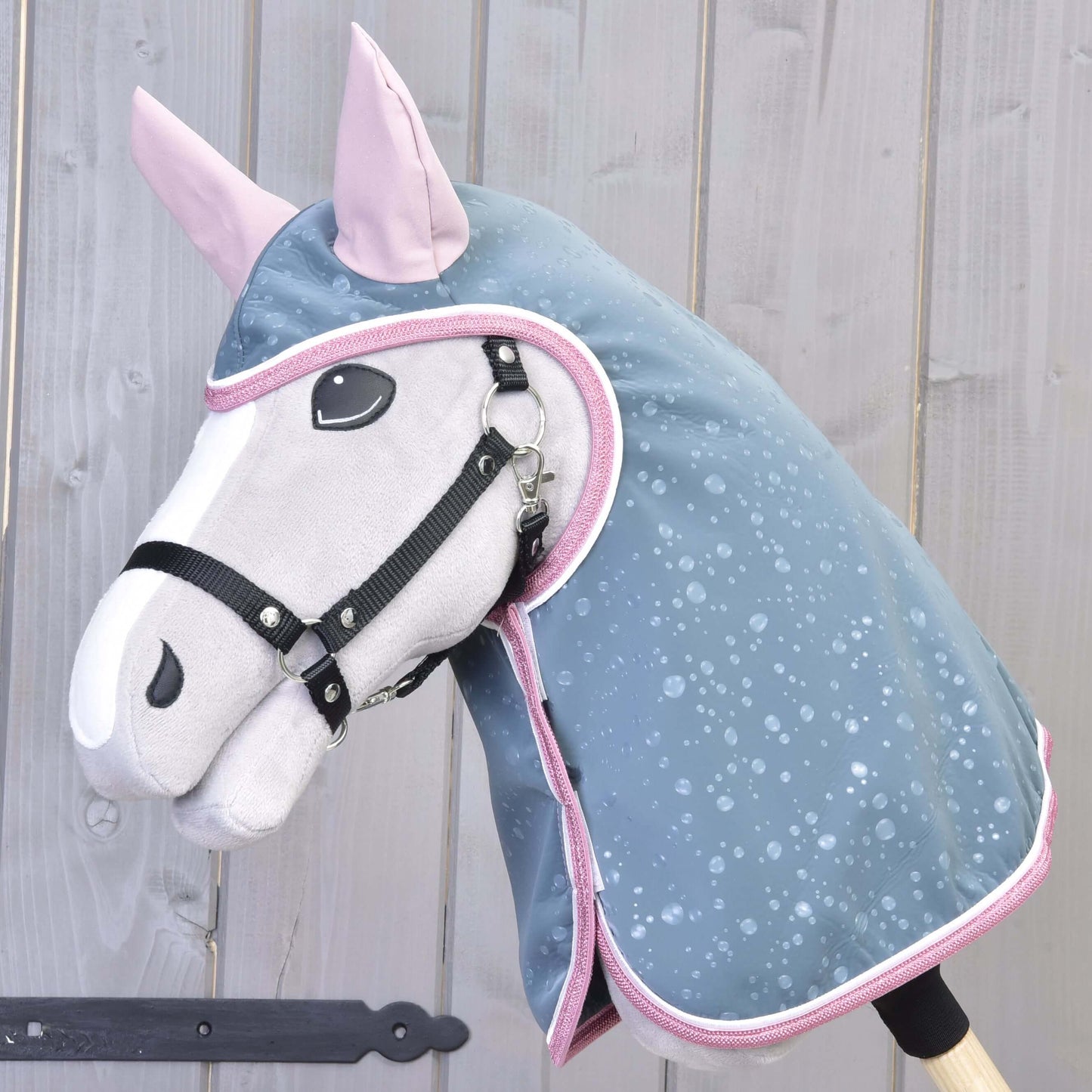 Hobby Horse LarDen „Pláštenka Best“ má plyšovú konskú hlavu s čiernou uzdečkou a modrou pláštenkou do každého počasia s ružovým lemovaním, nasadenú na drevenej tyči pred stenou zo svetlého dreva.