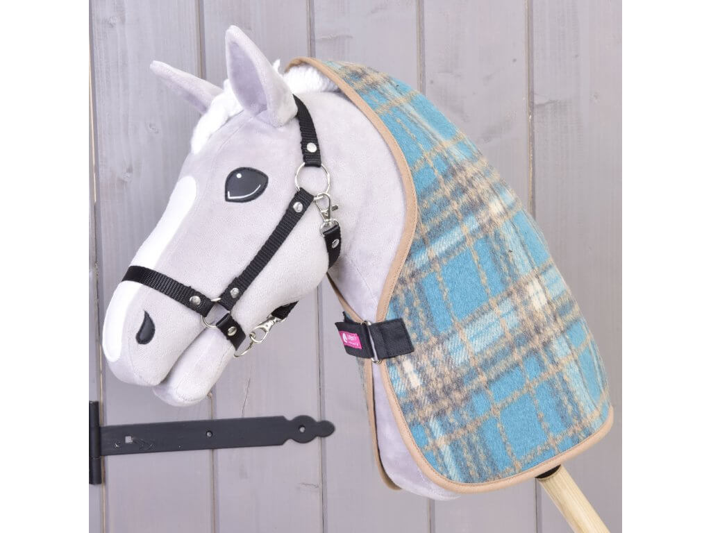 Plyšový Hobby Horse od Hobby Horse LarDen so sivo-bielou tvárou, čiernou uzdečkou a dekou Deka stajňová Scottish Cube v modrej kocke stojí na tyči pred svetlou drevenou stenou.