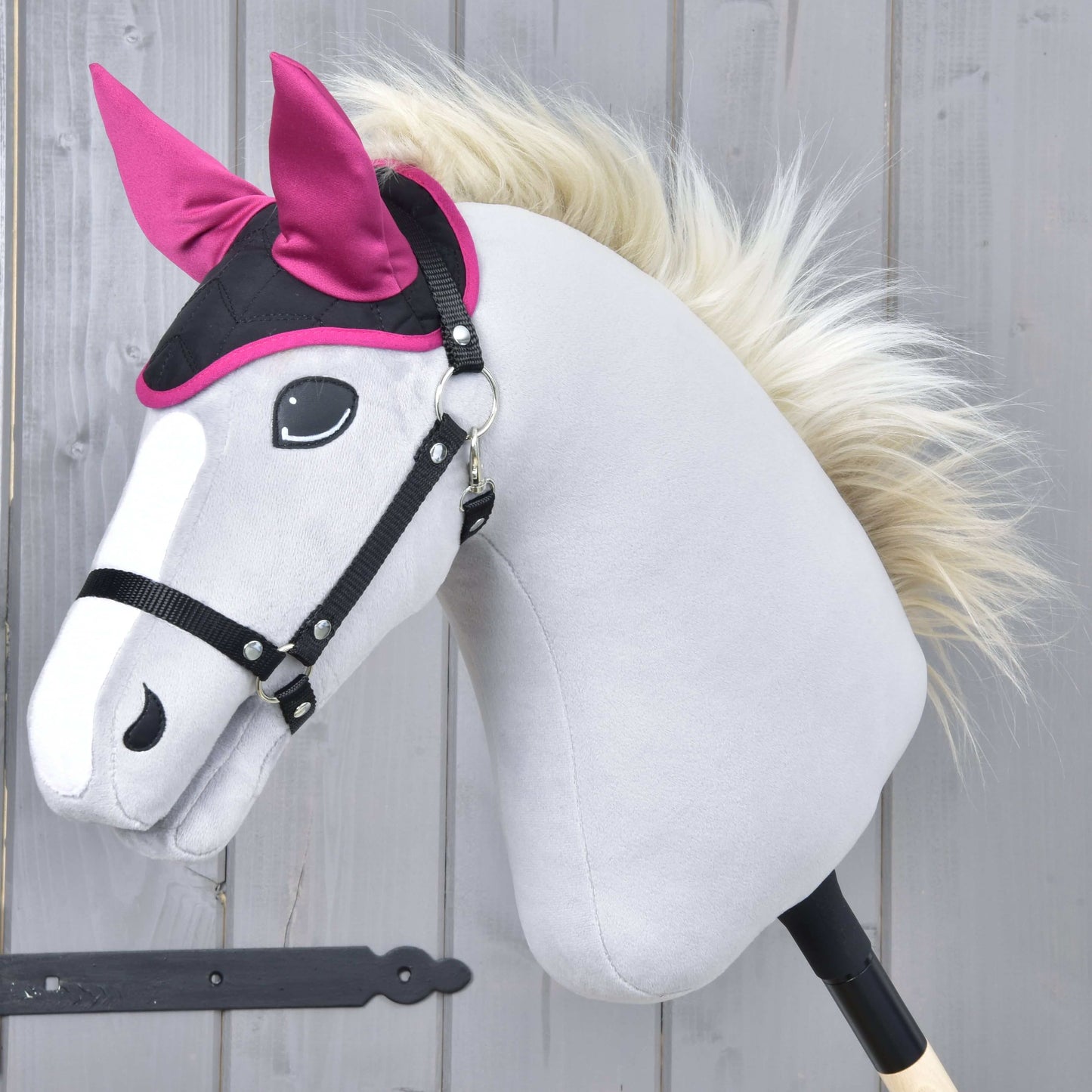 Sivá hračka Hobby Horse od Hobby Horse LarDen s čiernou uzdečkou, bielou hrivou a jasne ružovou čabrakou Thermo Sky, navrhnutou na ochranu pred hmyzom, pózuje pred pozadím z drevených dosiek.