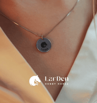 Detail osoby v krémovom oblečení s retiazkou s medailónikom LarDen z chirurgickej ocele od Hobby Horse LarDen s okrúhlym príveskom z chirurgickej nehrdzavejúcej ocele vyrytým konskou hlavou a nápisom LarDen. „Hobby Horse LarDen“ vidno v spodnej časti obrázka.