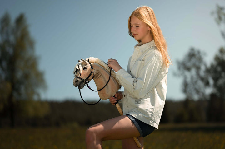 Mladý človek s dlhými blond vlasmi, vo svetlej bunde a kraťasoch, si užíva Hobby Horsing na Hobby Horse vonku na slnečnej trávnatej lúke so stromami v pozadí.