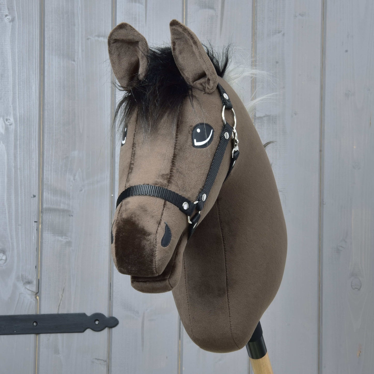Hobby Horse Alfredo s čiernou ohlávkou od Hobby Horse LarDen má plyšovú hnedú hlavu žrebca anglického plnokrvníka s čiernou hrivou a uzdečkou, je nasadený na drevenej tyči a umiestnený pred stenou zo sivých drevených panelov.