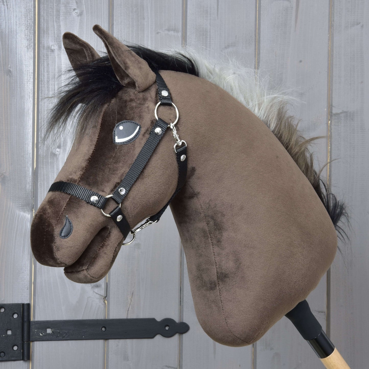 Hobby Horse Alfredo s čiernou ohlávkou od Hobby Horse LarDen má hnedú plyšovú hlavu v štýle plnokrvníka, čiernu hrivu, bielu ofinu a čiernu ohlávku, je nasadený na drevenej tyči a umiestnený pred sivým dreveným pozadím.