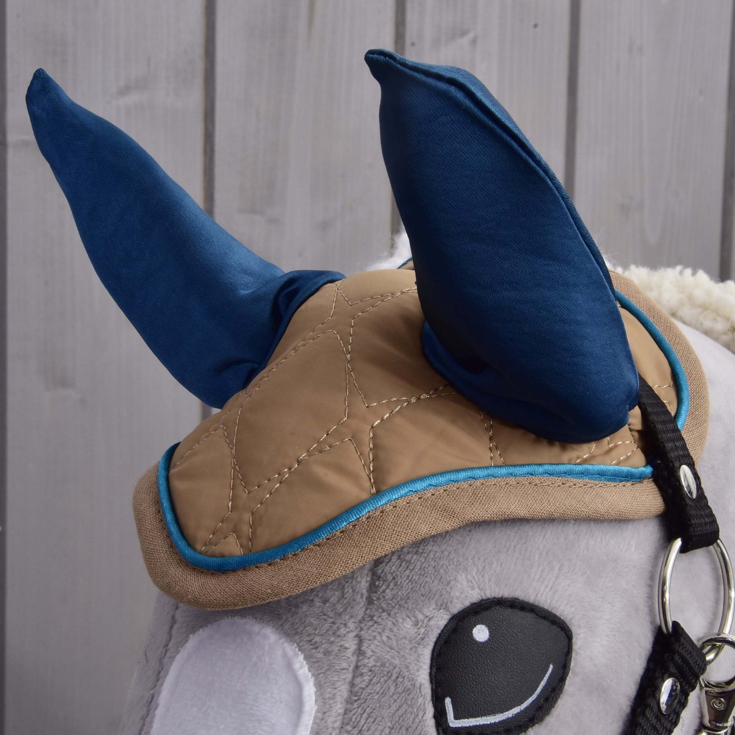 Detailní záběr na Set Almond Star od Hobby Horse LarDen ukazuje modrou čabraku, béžovou prošívanou deku s modrým lemováním a uzdečku na plyšové hlavě Hobby Horse na světlém dřevěném pozadí.