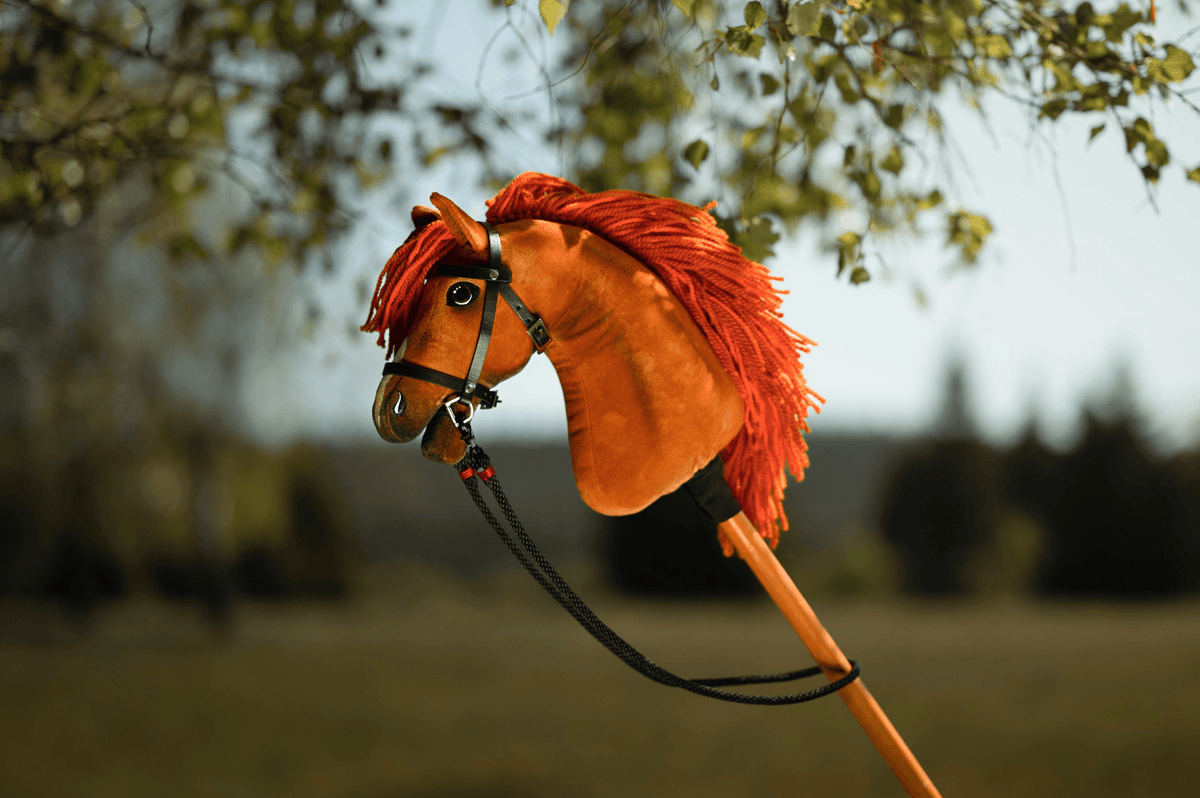 Plyšový Hobby Horse s oranžovou hrivou a uzdečkou je držaný vonku, ideálny pre dobrodružstvá pri Hobby Horsingu, so zelenými listami nad ním a jemne rozmazaným prírodným pozadím.