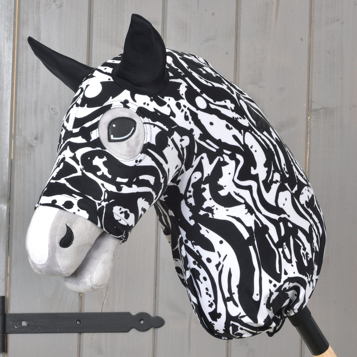 The Hobby Horse LarDen Hobby Horse protective suit Black & White, featuring a black and white design with cartoon-like eyes and black ears, is displayed mounted on a wooden stick against a gray wooden wall.