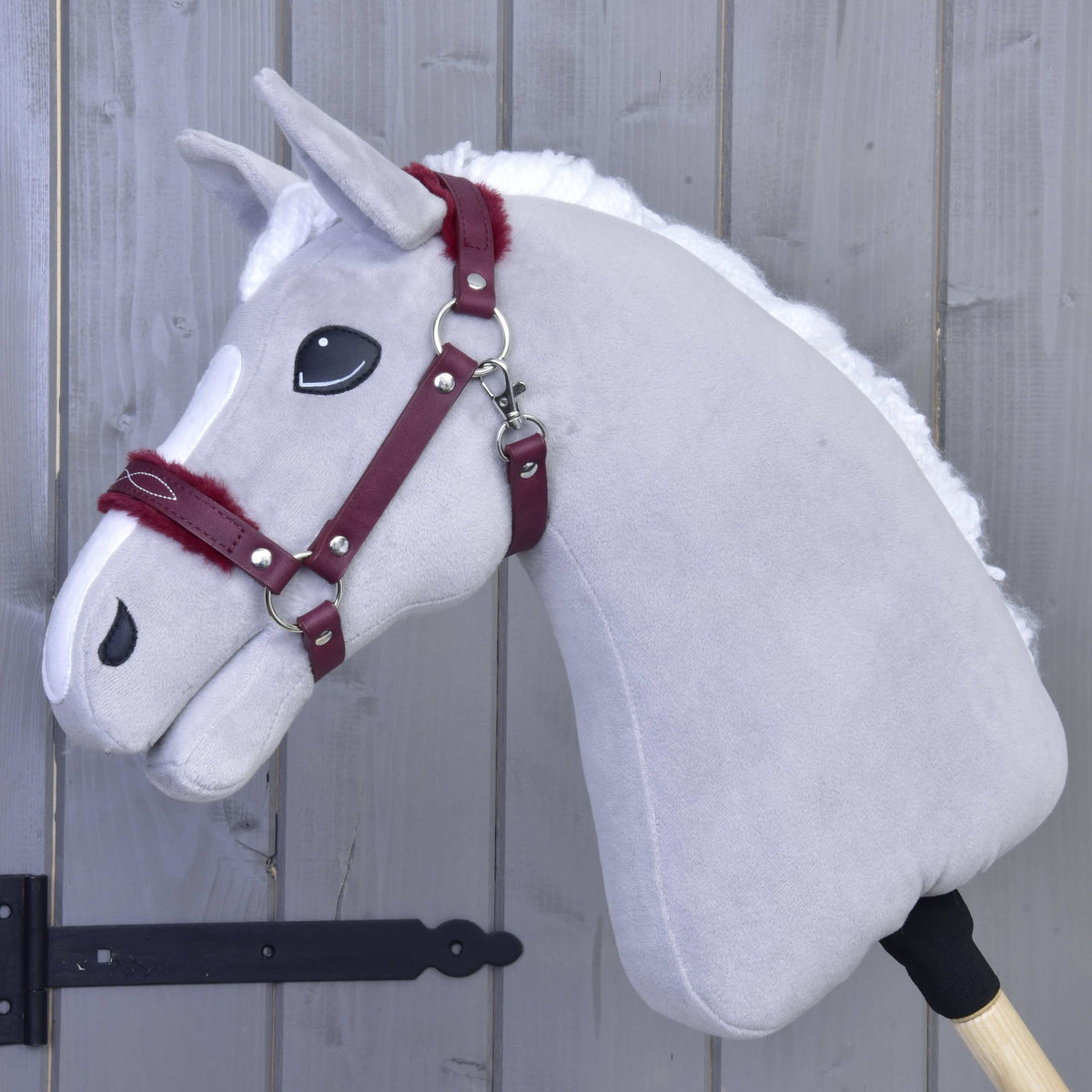 Ohlávka s barančekom z koženky Burgundy od Hobby Horse LarDen na sivom plyšovom Hobby Horse s bielou hrivou a vínovou uzdečkou dodáva štýlový vzhľad na pozadí sivej drevenej steny — je tak dokonalým doplnkom.