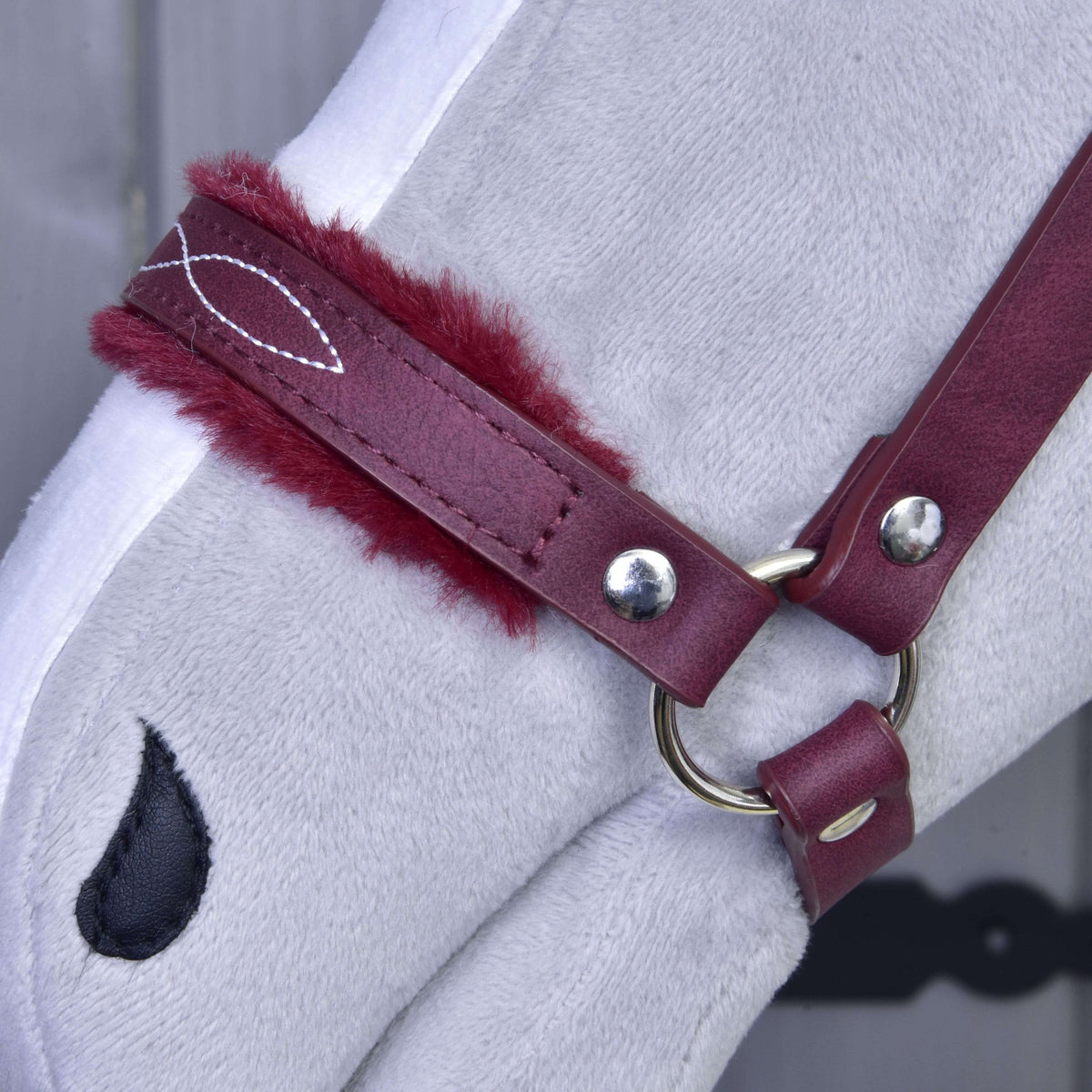 Detail hlavy svetlosivého plyšového koňa s Hobby Horse LarDen „Ohlávka s barančekom z koženky Burgundy“. Obsahuje vínovo polstrovanú červenú koženku a čiernu kvapkovitú nášivku nozdier. Ideálna ako štýlový doplnok pre Hobby Horse. Pozadie je rozostrené.