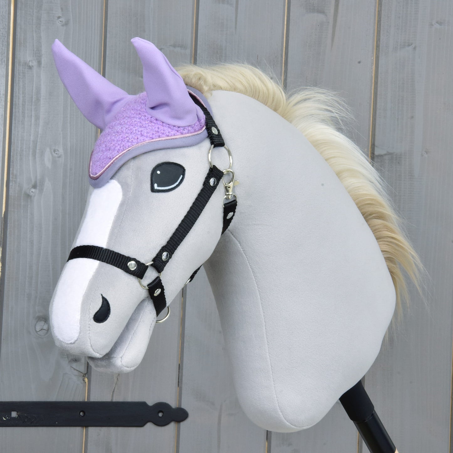 Plyšový Hobby Horse so svetlosivým telom, čiernou uzdečkou a krémovou hrivou nosí Čabraka Orchid od Hobby Horse LarDen pre ochranu pred hmyzom. V pozadí je drevený plot.