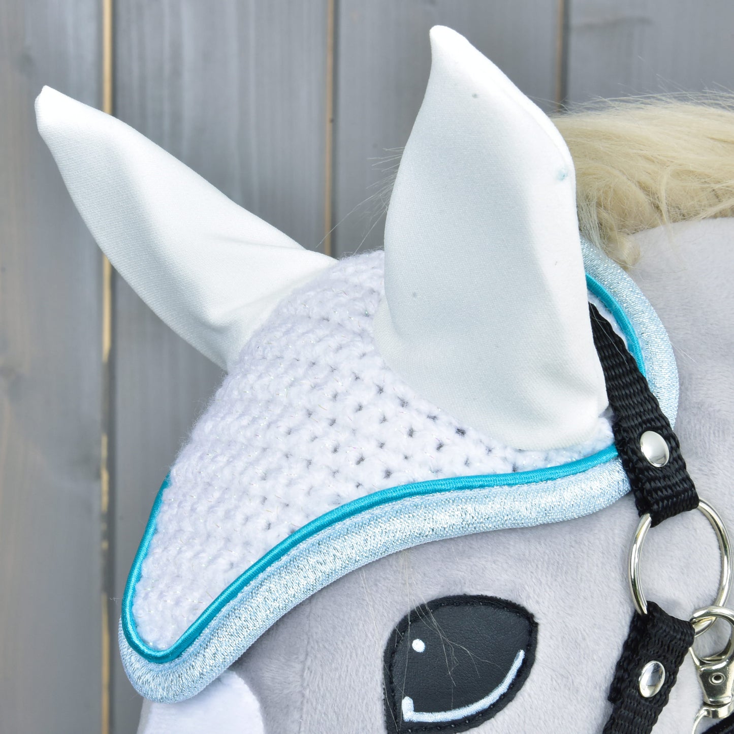 Detail sivého plyšového koňa s čabrakou háčkovanou Glitter Cuties od Hobby Horse LarDen, s modrým lemom, dvoma vztýčenými bielymi ušami a čiernou uzdečkou, na svetlo drevenom pozadí.