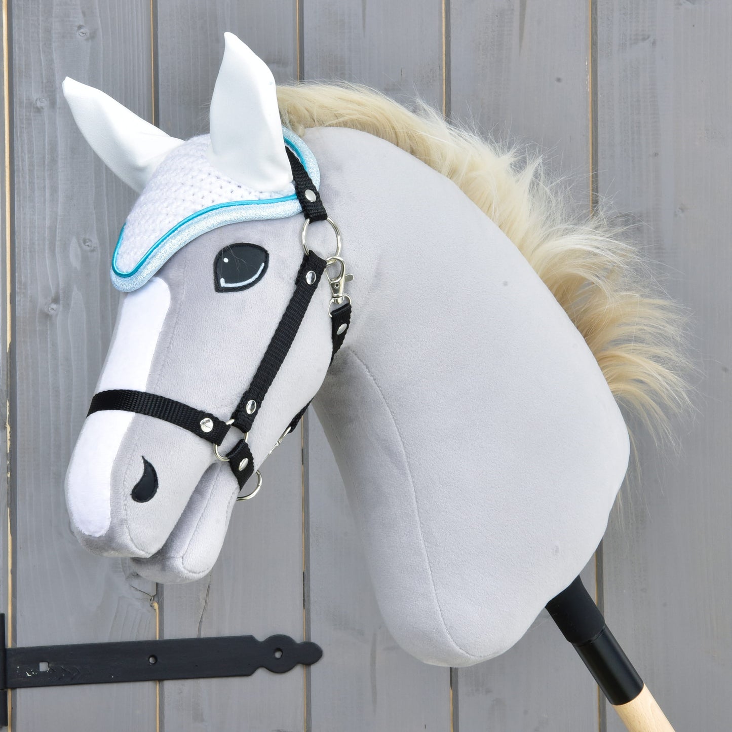 Detail sivého plyšového Hobby Horse od Hobby Horse LarDen s bielou hrivou, čiernou uzdečkou a čabrakou háčkovanou Glitter Cuties pre ochranu proti muchám, nasadeného na drevenej tyči pred dreveným plotom.