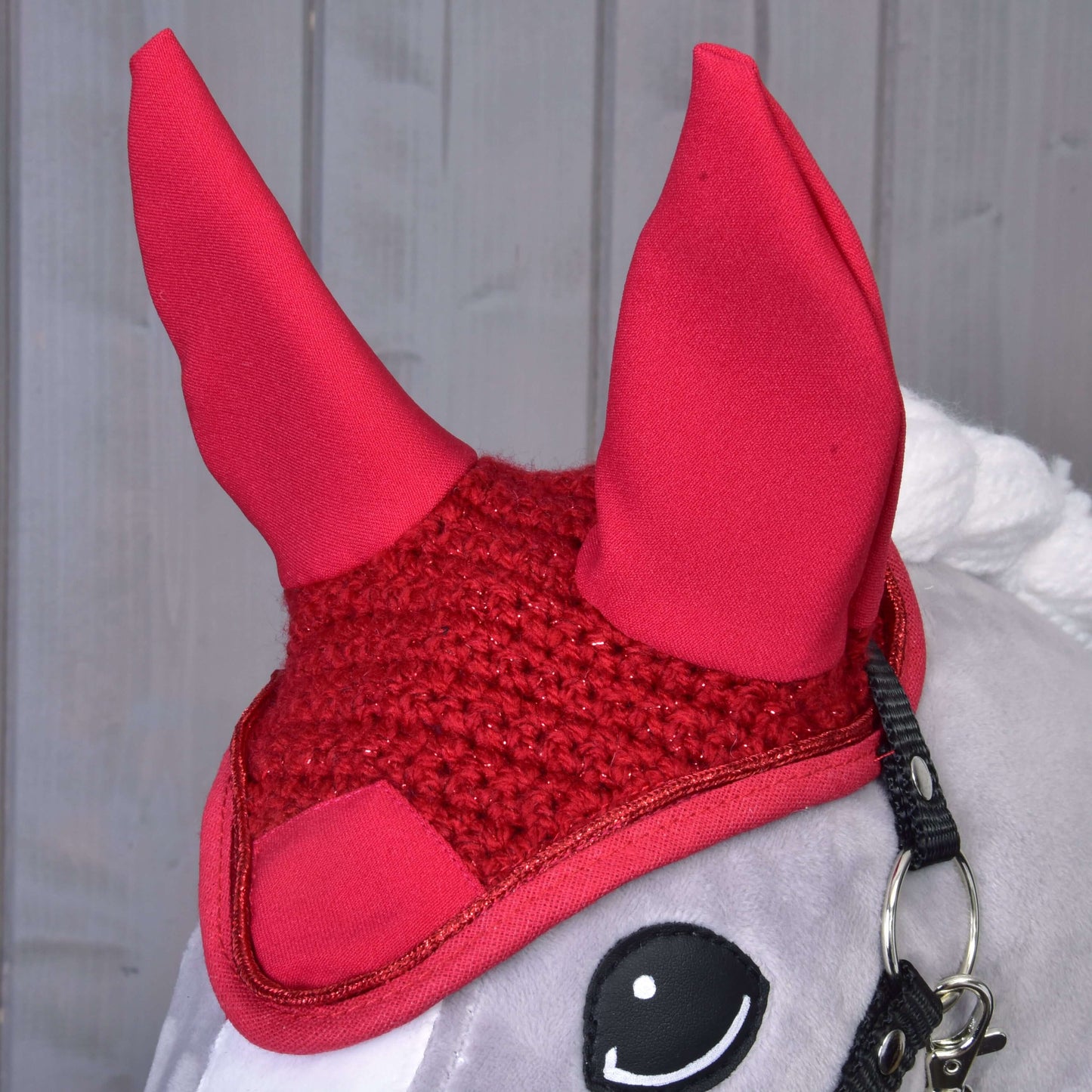 Detail sivého plyšového koňa s čabrakou Hobby Horse LarDen Čabraka háčkovaná červená s vysokými špicatými ušami, poskytujúcou štýlovú ochranu proti muchám, pred svetlým dreveným pozadím.