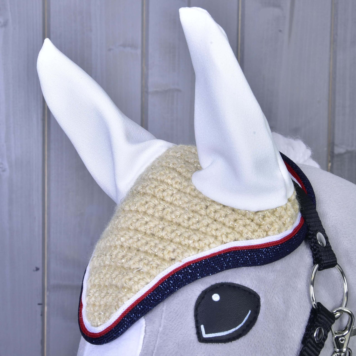Detail hlavy koně s čabrakou Nicol od Hobby Horse LarDen v béžové barvě s bílýma ušima, modrým a červeným lemováním a černou ohlávkou, na pozadí šedého dřeva.