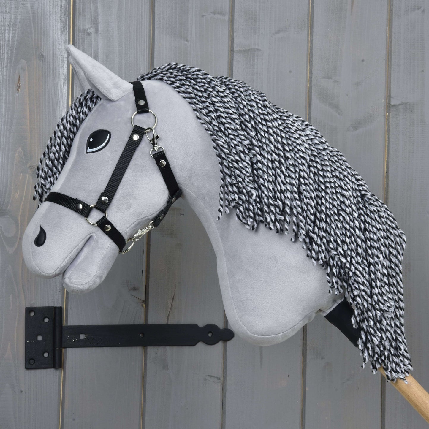 Hobby Horse Caesar s čiernou ohlávkou od Hobby Horse LarDen, so sivou plyšovou hlavou, čiernobielou priadzovou hrivou, čiernou ohlávkou a veľkými kreslenými očami na drevenej tyči – ideálny pre nápaditú hru.