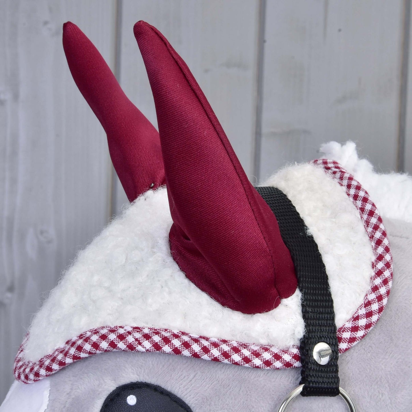 Detail plyšového zvieratka s Čabrakou Cube Red od Hobby Horse LarDen, s červenými látkovými rohmi, červenobielym károvaným lemom a čiernym pásikom s kovovým krúžkom, na sivom drevenom pozadí.