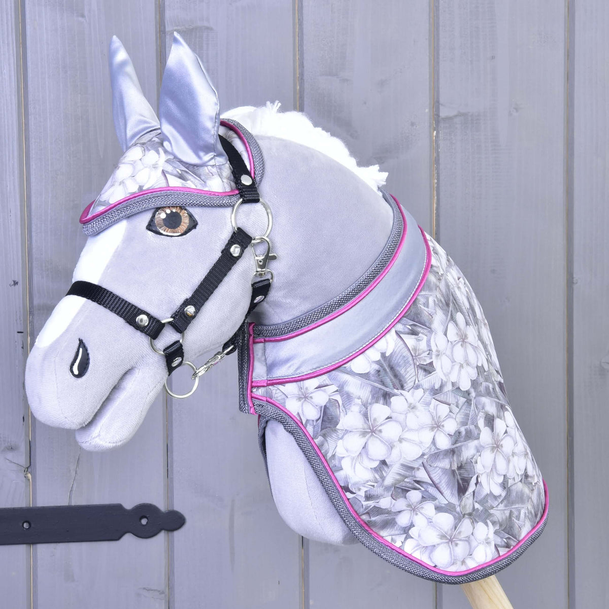 Set Grey Flower od Hobby Horse LarDen na sivom plyšovom Hobby Horse s čiernou uzdečkou, strieborno-ružovým vybavením, kvetinovým vzorom a ladiacou dečkou, na pozadí zo svetlosivého dreva.