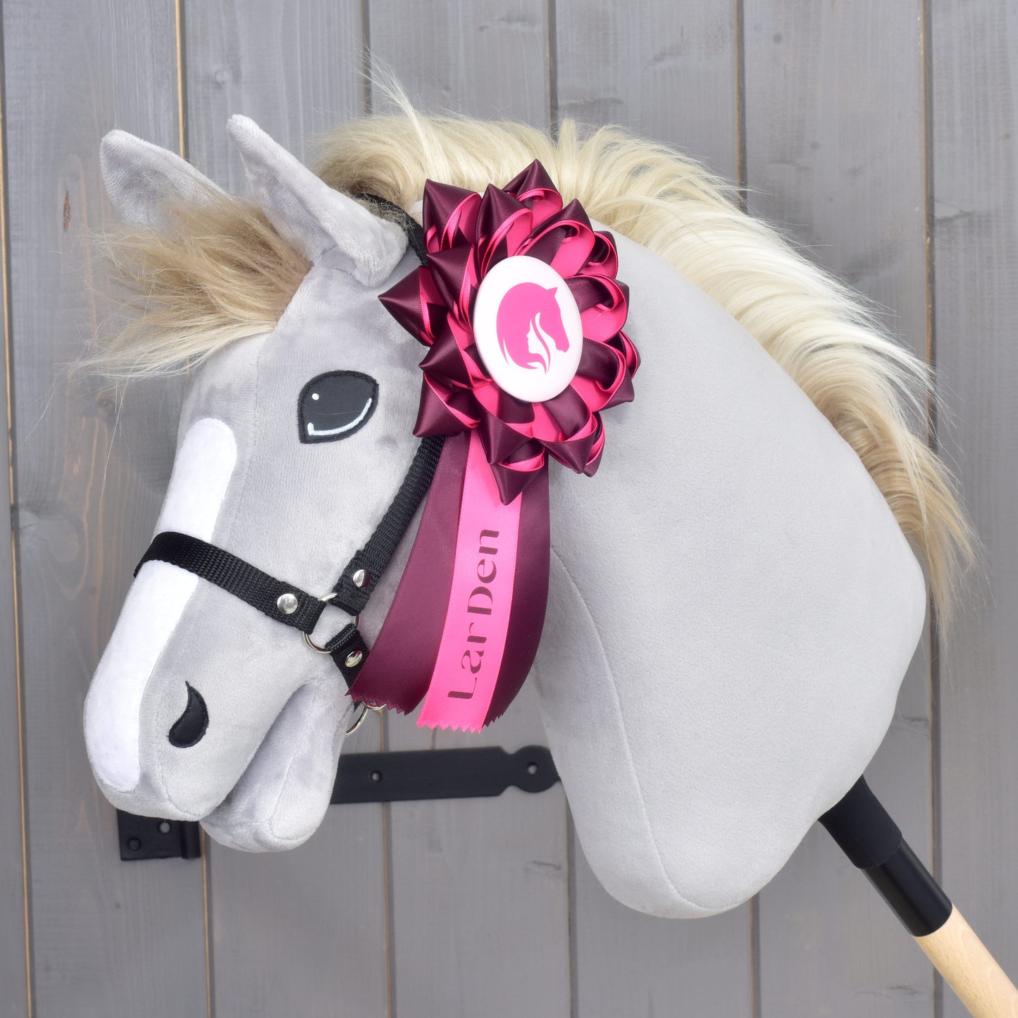 Plyšový Hobby Horse Kokarda Raspberry Rose od Hobby Horse LarDen so sivou tvárou, krémovou hrivou a čiernou uzdečkou na tyči, ozdobený veľkou ružovo-bielou drezúrnou kokardou s logom LarDen na krku. Pozadie tvorí drevené obloženie.