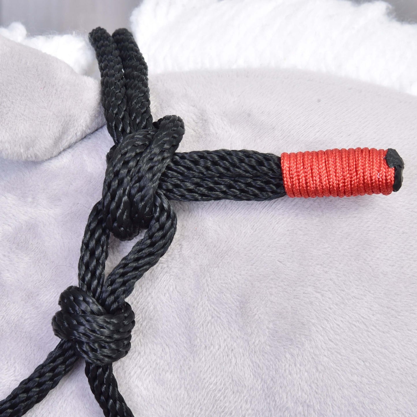 Detail čierneho polypropylénového Povrazový set Spark od Hobby Horse LarDen s dvoma uzlami na svetlosivej látke. Jeden koniec je úhľadne omotaný jasne červenou niťou, ktorá zvýrazňuje jedinečný dizajn tohto ručne vyrábaného doplnku.