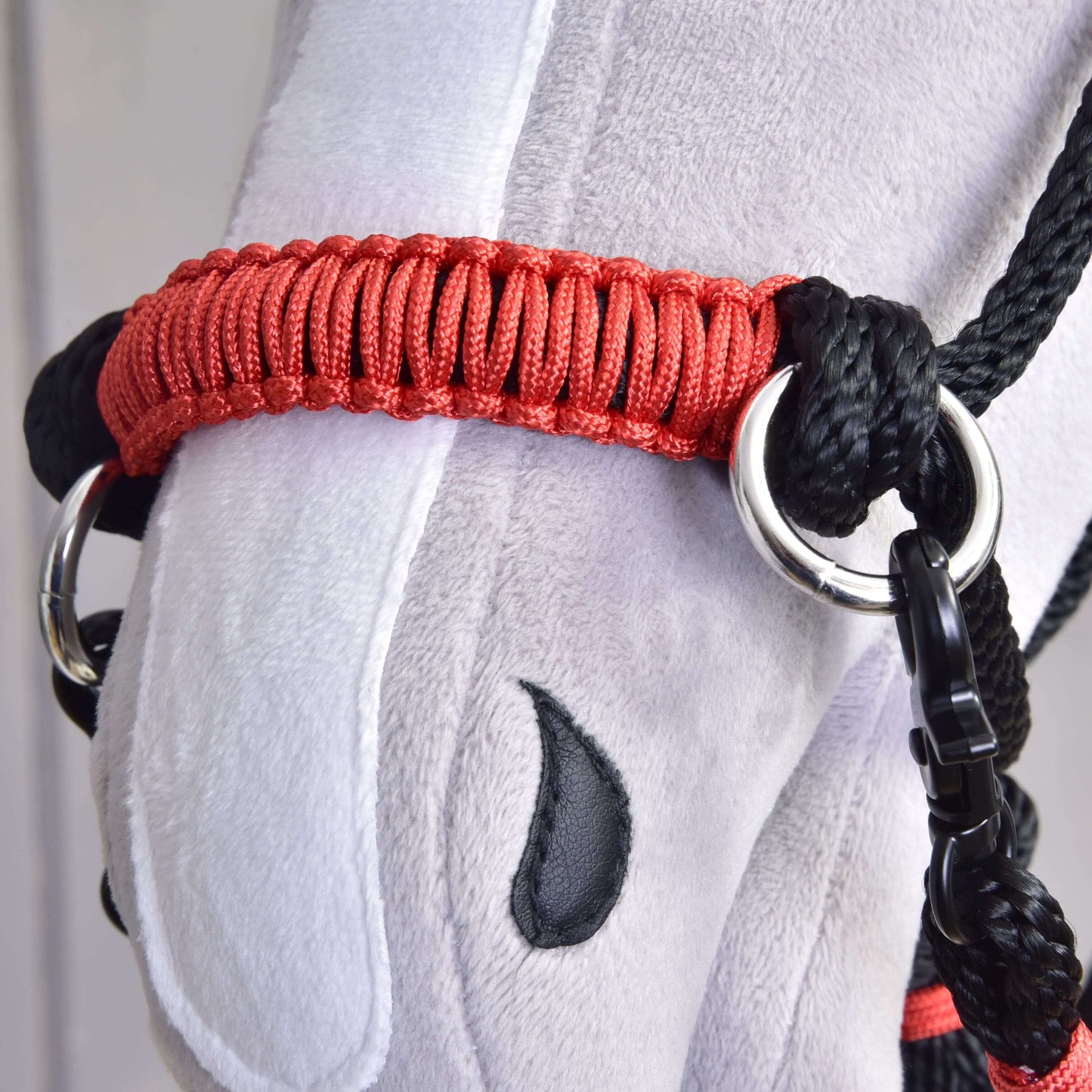 Detail hlavy plyšového koňa s červenou a čiernou ohlávkou, čiernym vyšívaným okom a sivo-bielou látkou, s Hobby Horse LarDen Povrazový set Spark, vyrobeným z odolného polypropylénu ako jedinečný ručne vyrábaný doplnok.