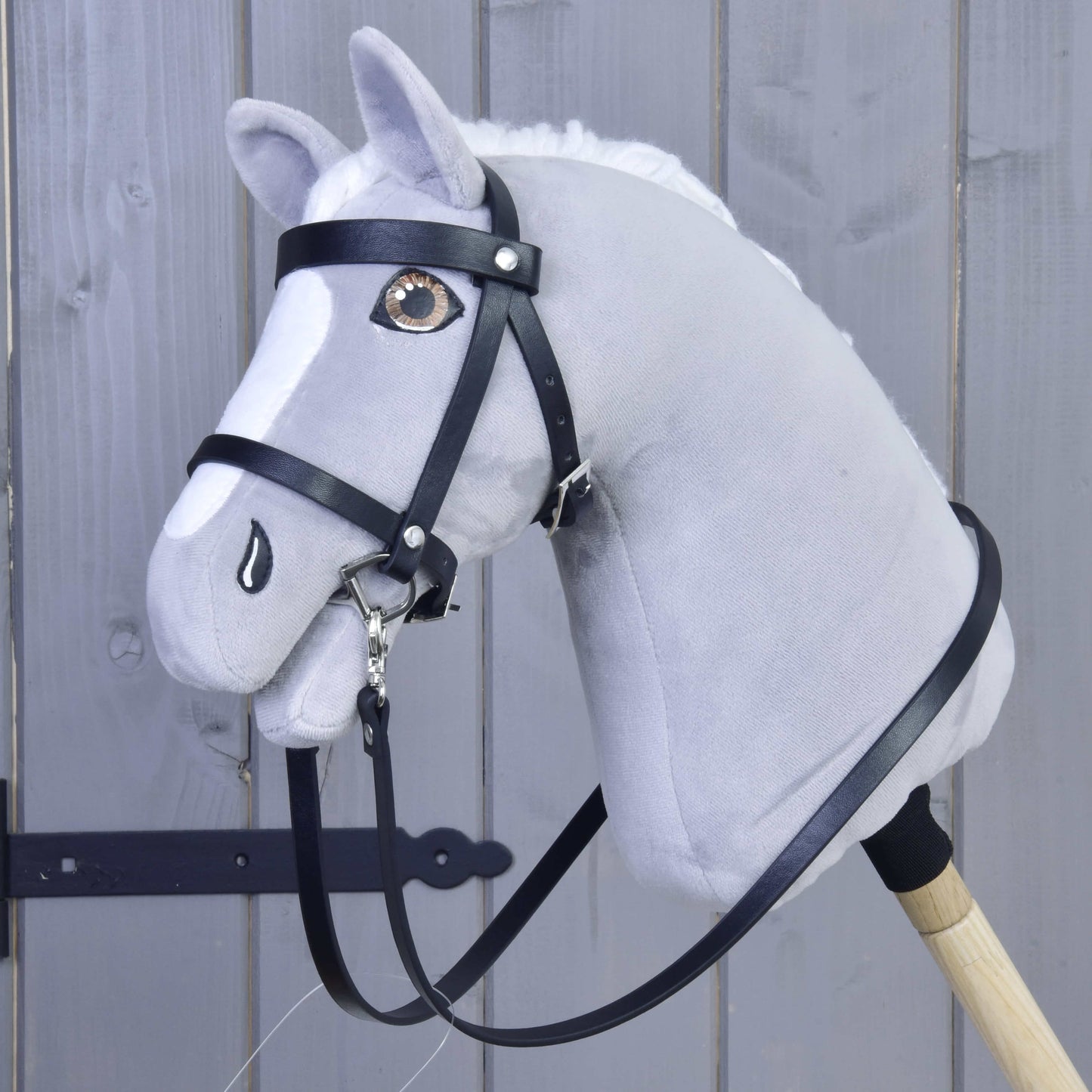 Hobby Horse LarDen Set Basic, sivý plyšový Hobby Horse s čiernou uzdečkou a opratami na drevenej tyči, je zobrazený pred sivými drevenými panelmi.