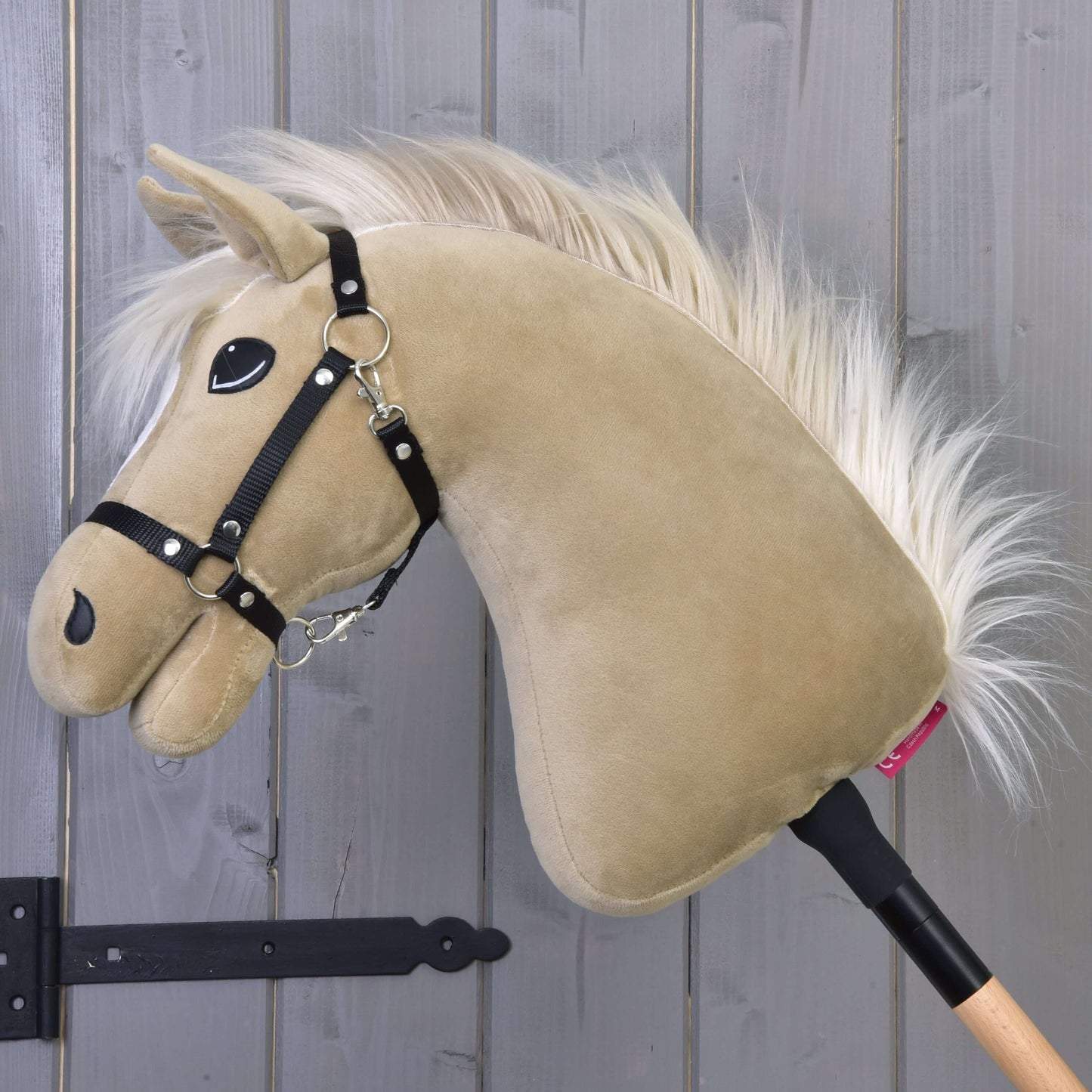 Hobby Horse Dancing Lady s čiernou ohlávkou od Hobby Horse LarDen s plyšovou hlavou kobyly anglického plnokrvníka v béžovej farbe a s bielou hrivou na drevenej tyči, zobrazený pred sivou stenou z drevených panelov.