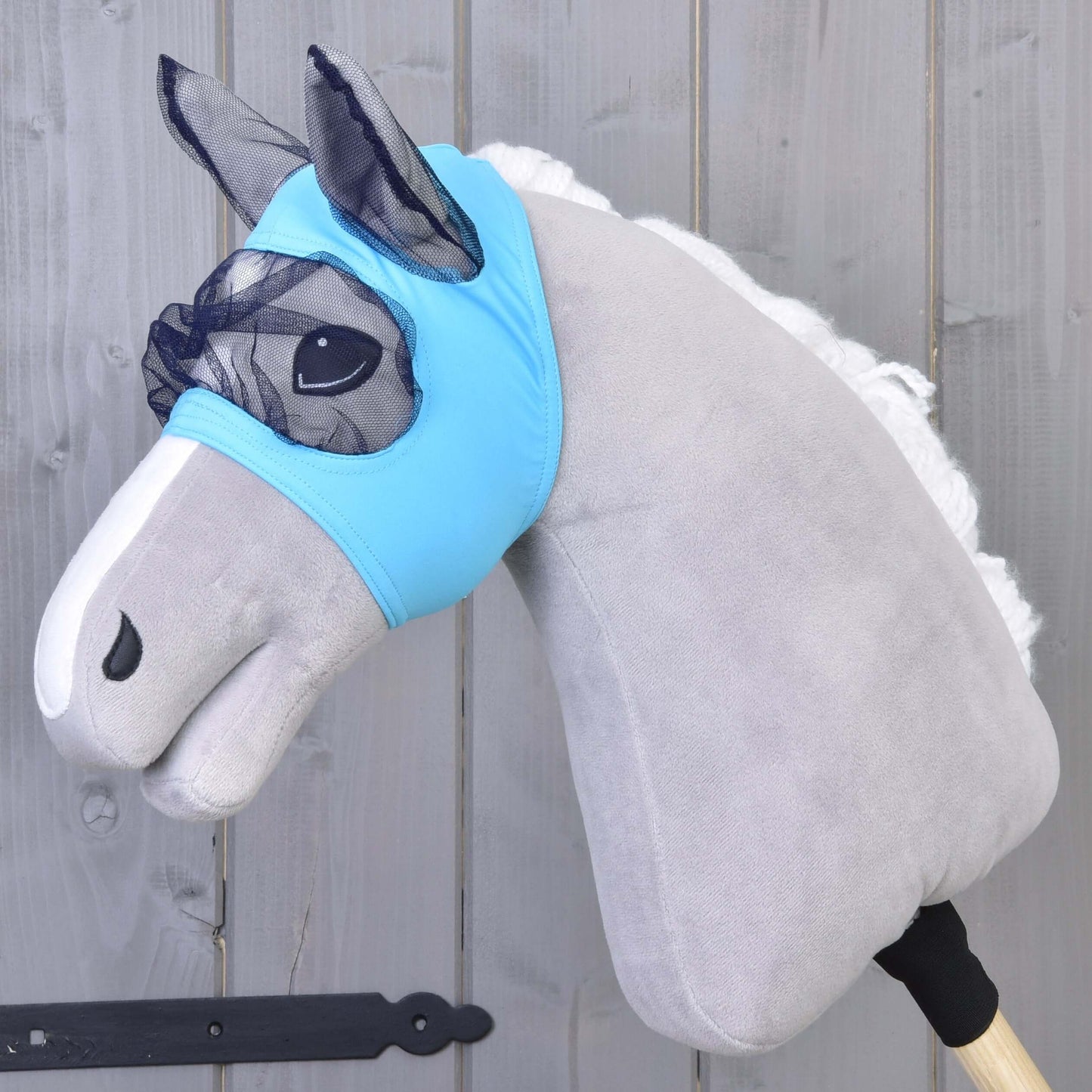 Plyšový Hobby Horse so svetlosivým telom, bielou hrivou a modrou prateľnou Maska proti hmyzu od Hobby Horse LarDen, nasadený na drevenej tyči na pozadí sivého dreva.
