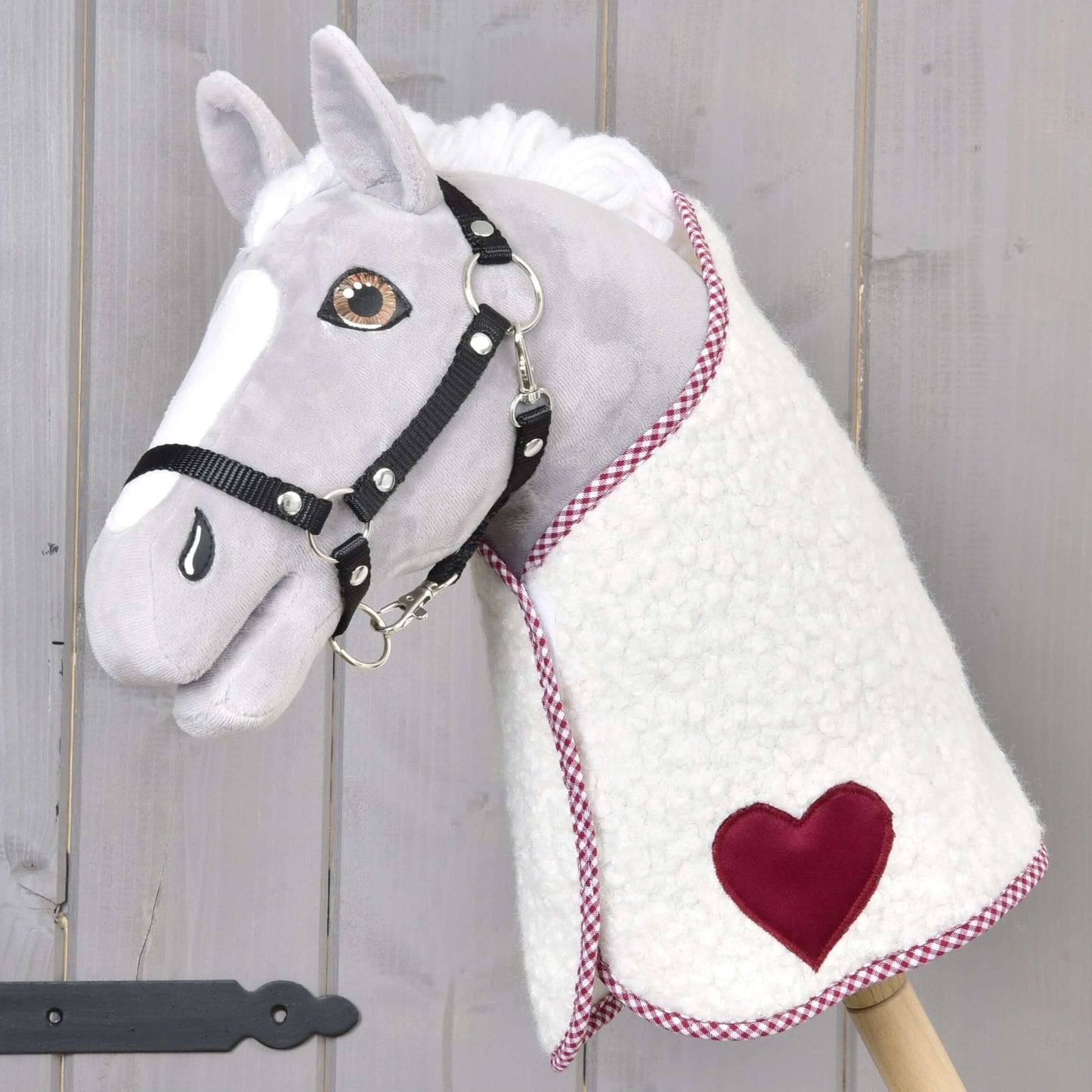 Plyšový Hobby Horse od Hobby Horse LarDen s bielosivou tvárou, čiernou uzdečkou a fleecovou Dekou Cube Red s červenou nášivkou v tvare srdca, nasadený na drevenej tyči na pozadí svetlého dreva.