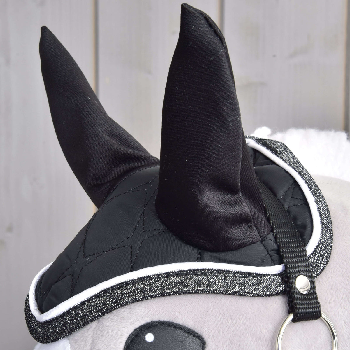 Detail Set Thermo Black Queen od Hobby Horse LarDen s plyšovými čiernymi ušami s čabrakou, prešívaným krytím s trblietavým lemom ako deka pre Hobby Horse a pásikom s kovovým krúžkom, pred svetlým dreveným obložením.