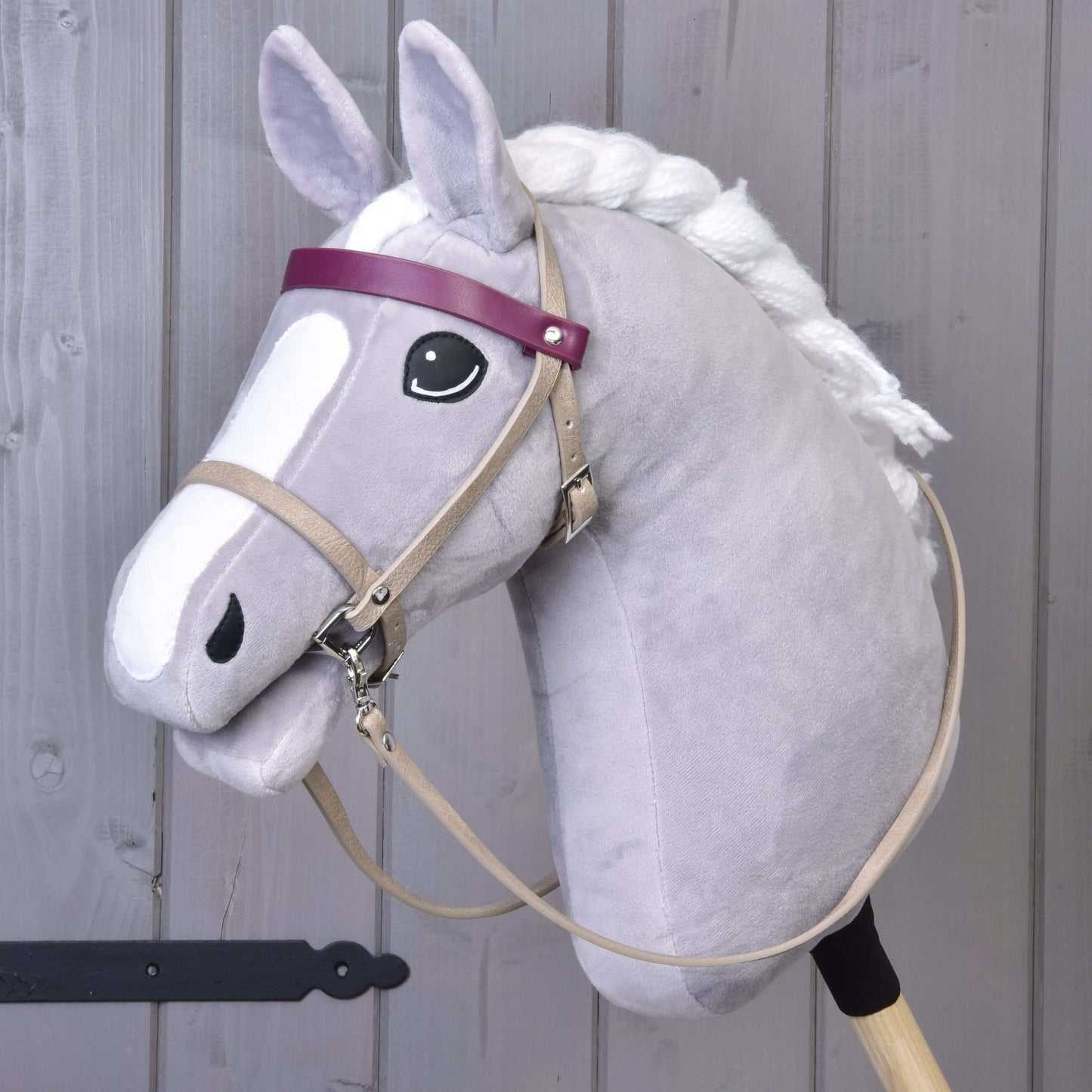 Hobby Horse LarDen so sivým Hobby Horse s bielou hrivou, čiernym okom a koženkovým setom Bordeaux – uzdečka, opraty a martingal –, doplneným o fialovú čelenku a tvár v kreslenom štýle. Stojí vzpriamene pred dreveným pozadím.