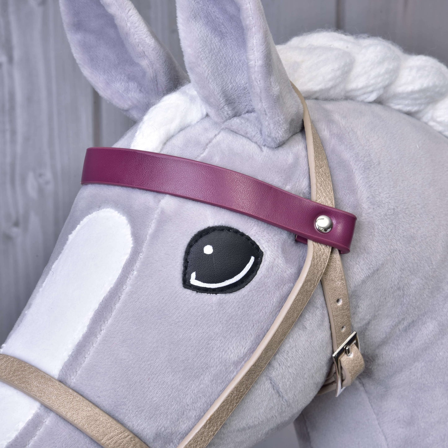 Detail Hobby Horse LarDen s bielou hrivou a vyšitým čiernym okom, ktorý má na sebe koženkový set Bordeaux – uzdečka, opraty a martingal. Má fialovú čelenku a je vyobrazený pred dreveným pozadím.