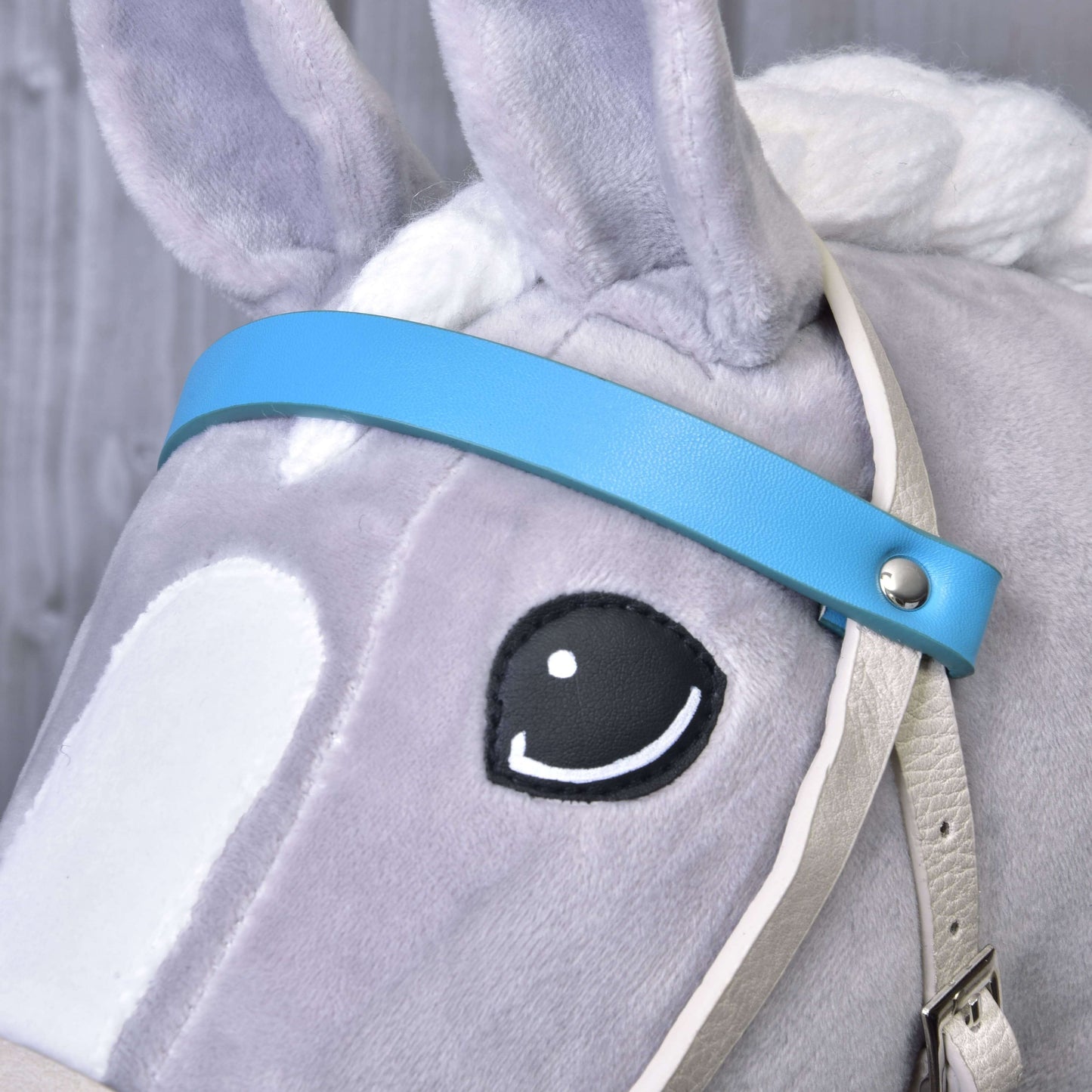 Detail šedého plyšového Hobby Horse od Hobby Horse LarDen s koženkovým setem Aqua – uzdečka, otěže a martingal –, bílou hřívou a jemným černým okem. Pozadí tvoří jemně rozmazané dřevěné obložení.