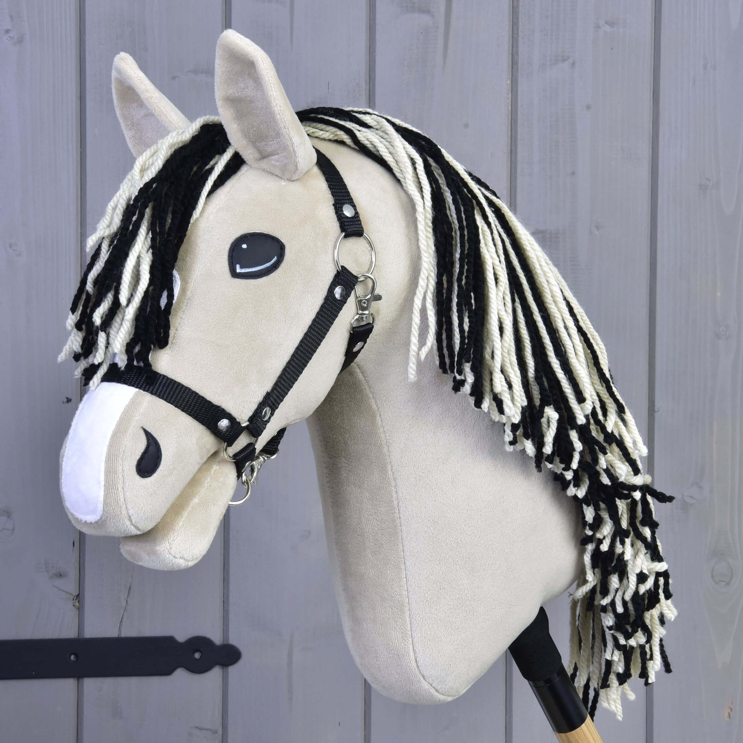 Hobby Horse Aurora s čiernou ohlávkou od Hobby Horse LarDen, s béžovou plyšovou hlavou, čierno-bielou priadzovou hrivou a na drevenej tyči, pred sivou drevenou stenou.