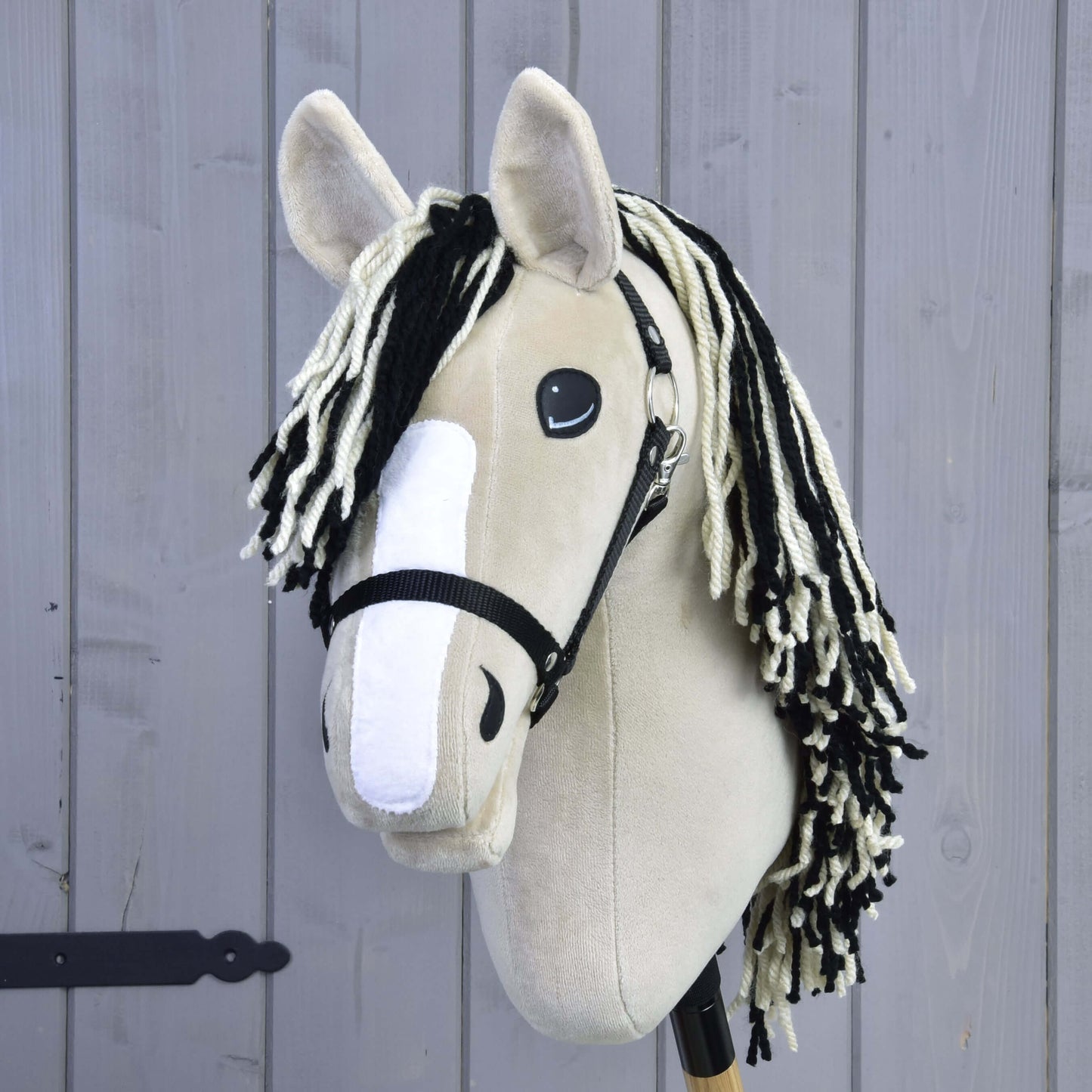 Hobby Horse Aurora s čiernou ohlávkou od Hobby Horse LarDen, s béžovým plyšovým prevedením inšpirovaným American Quarter Horse, čiernou ohlávkou, čierno-bielou priadzovou hrivou, bielym plsteným pruhom na nose a na drevenej tyči pred svetlosivou drevenou stenou.