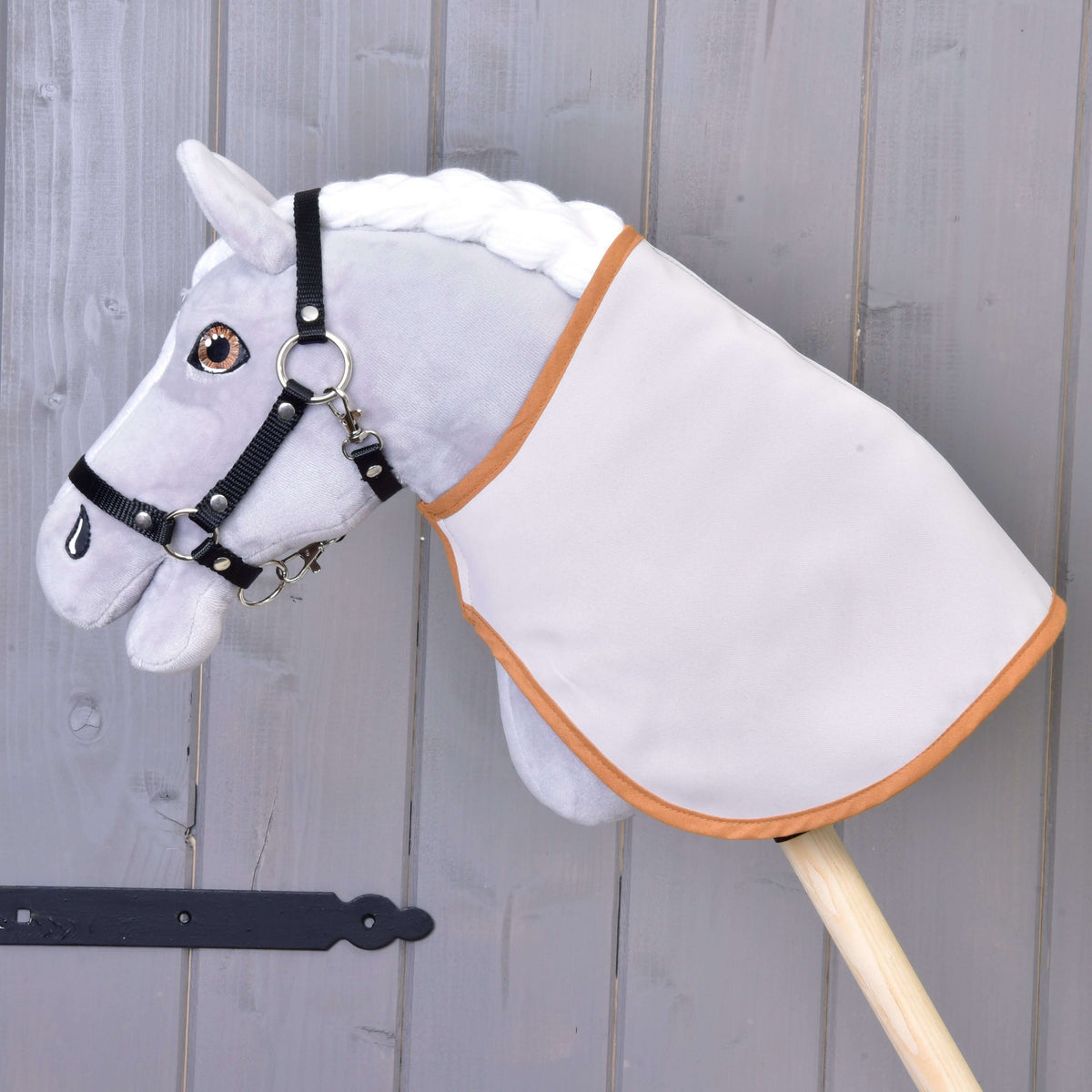 Sivý Hobby Horse LarDen s bielou hrivou, čiernou uzdečkou a Dekou anglická kocka, hnedá a obojstranná, nasadený na drevenej tyči pred sivou drevenou stenou.