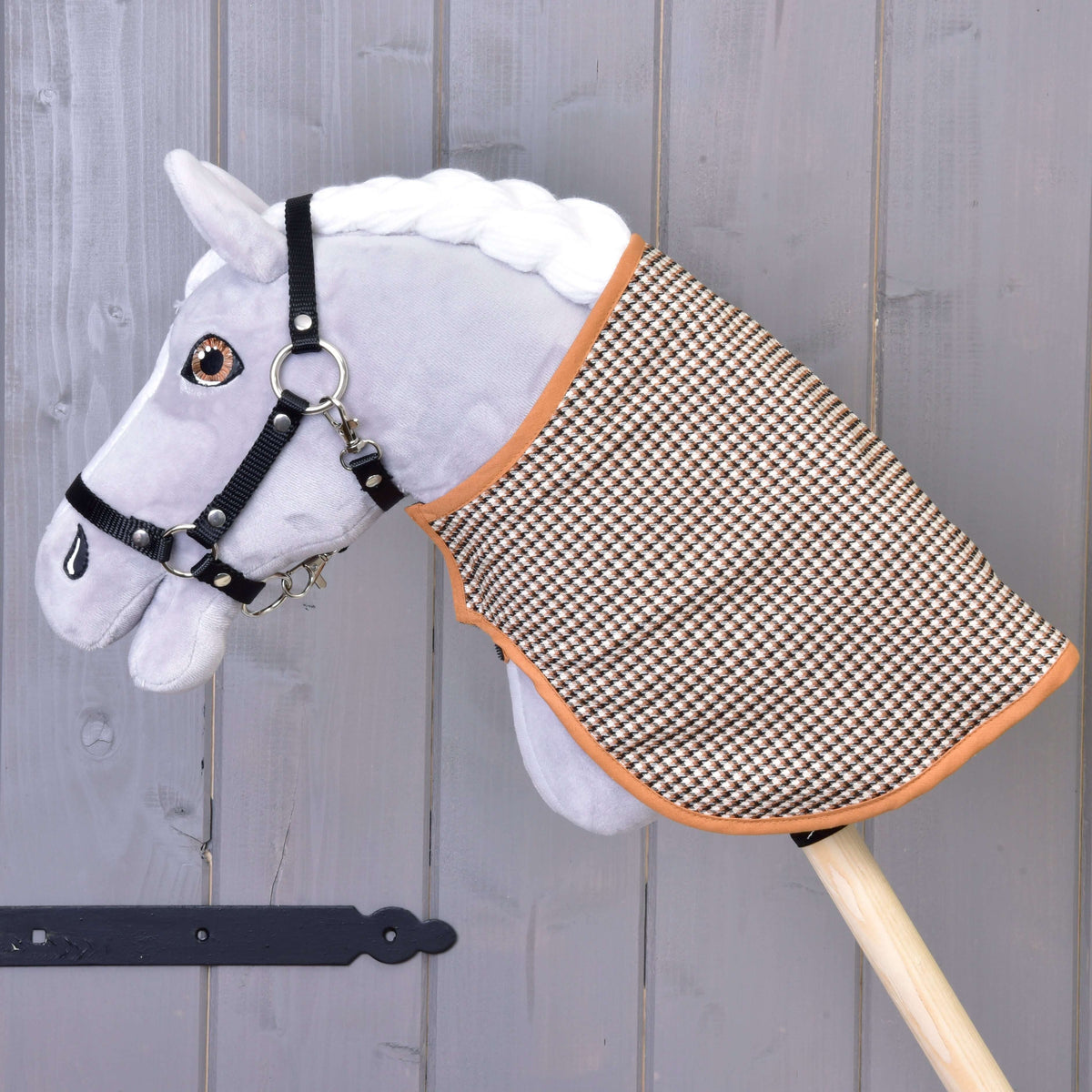 Plyšový Hobby Horse od Hobby Horse LarDen s bielou hrivou, čiernou uzdečkou a Dekou anglická kocka, hnedá a obojstranná, nasadený na drevenej tyči na pozadí sivej drevenej steny.