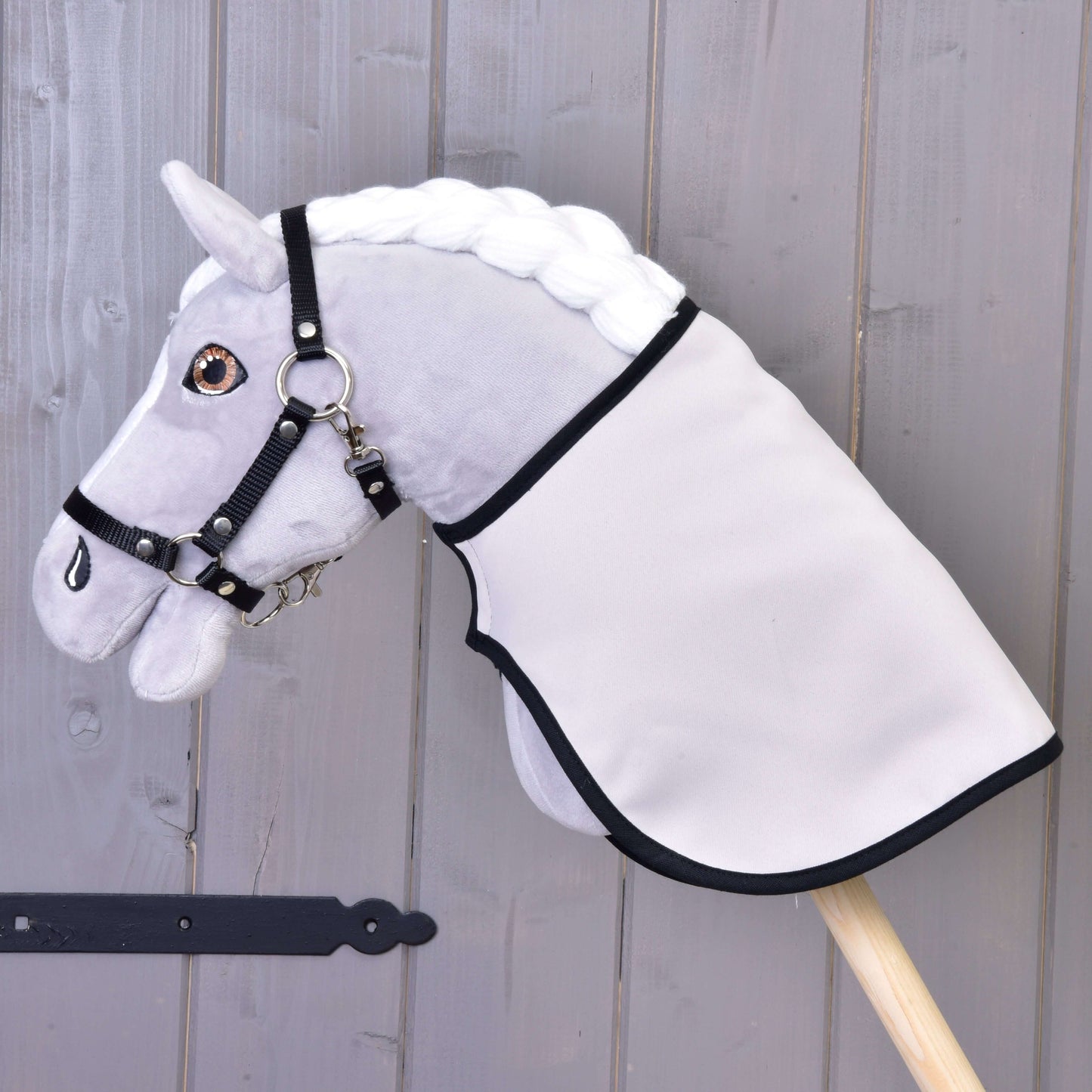 Sivý plyšový Hobby Horse s bielou hrivou, čiernou uzdečkou a Dekou anglická kocka čierna obojstranná od Hobby Horse LarDen, nasadený na drevenej tyči na pozadí steny z drevených dosiek.