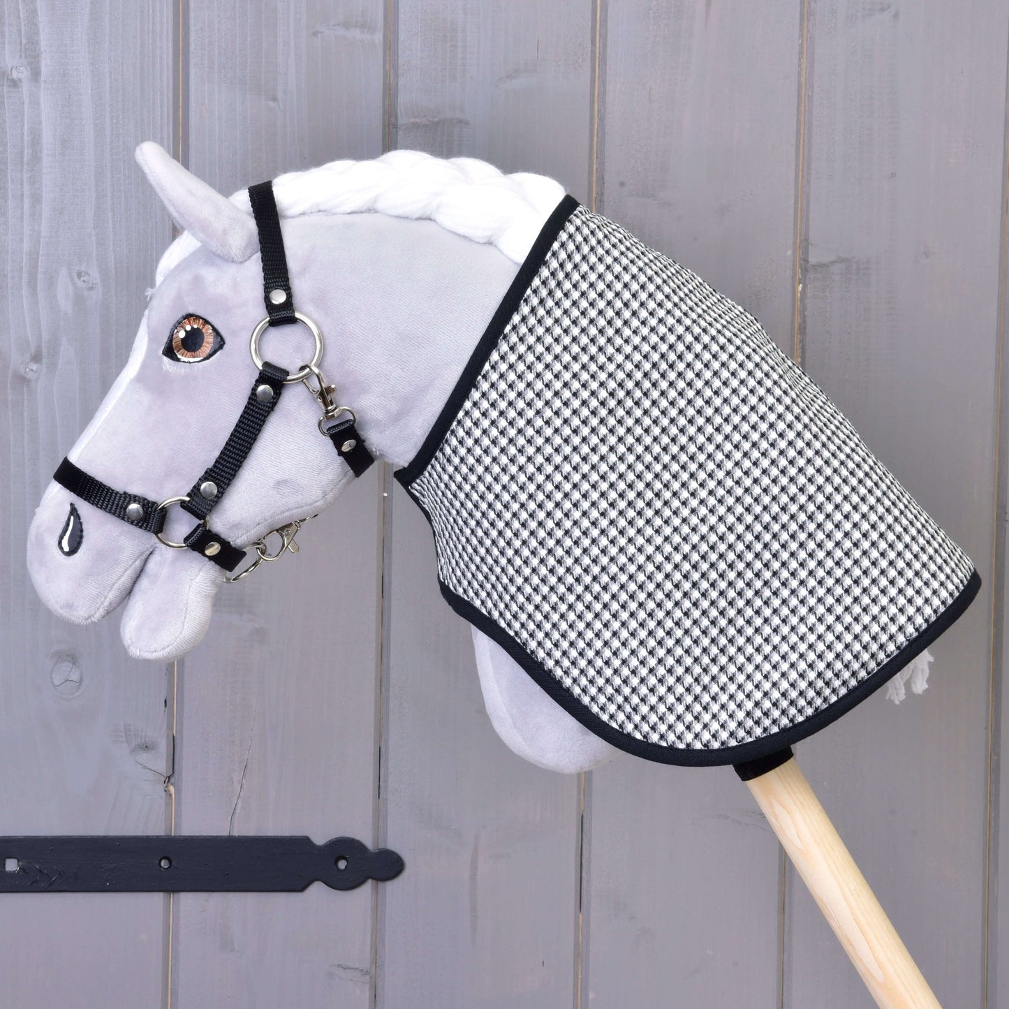 Sivý plyšový Hobby Horse s čiernou uzdečkou a Dekou anglická kocka čierna obojstranná od Hobby Horse LarDen, nasadený na drevenej tyči na pozadí sivej drevenej steny.