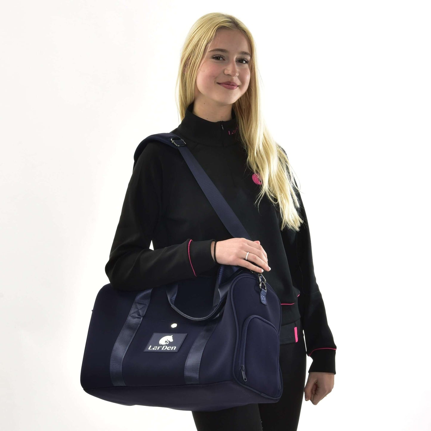 Usmievavá žena s dlhými blond vlasmi v čiernej bunde s ružovým lemovaním stojí na bielom pozadí a drží Športovú tašku LarDen od Hobby Horse LarDen, tmavomodrú tašku s nastaviteľným popruhom, ideálnu pre každodenné použitie.