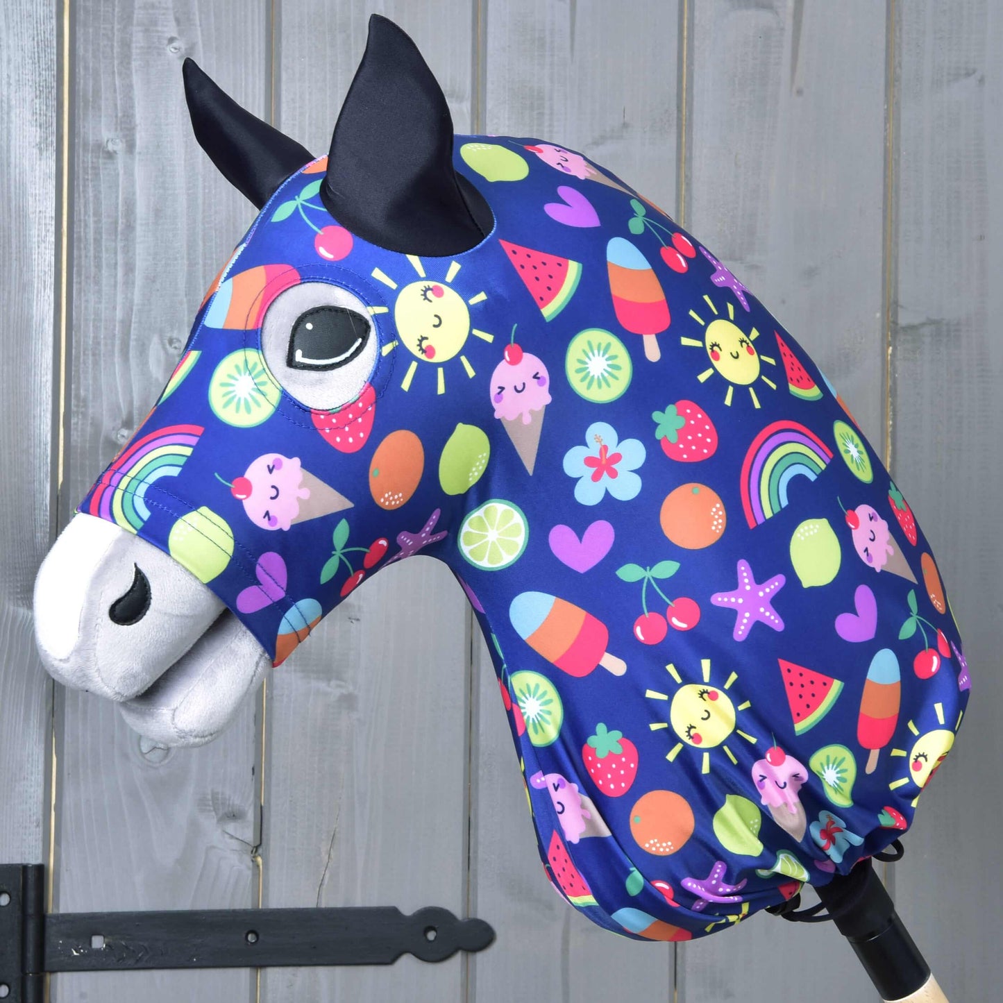 Látková hlava Hobby Horse s čiernymi ušami, veľkými očami a modrým ochranným oblekom Hobby Horse Happiness od Hobby Horse LarDen so slnkami, dúhami, srdiečkami, ovocím a zmrzlinou, na drevenom pozadí.