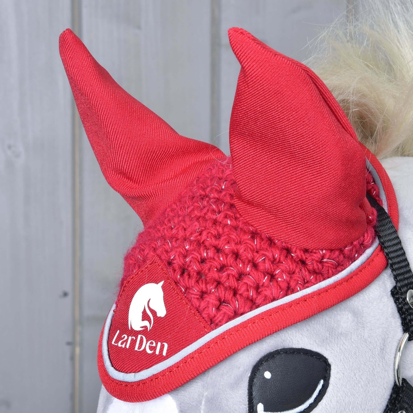 Detail koně s čabrakou Hobby Horse LarDen Čabraka háčkovaná Red sport s bílým logem LarDen a grafikou koně na pozadí světle šedých dřevěných panelů.