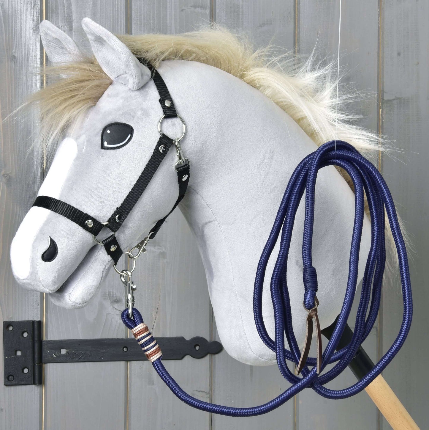 Hobby Horse - lonžovacie vodidlo od Hobby Horse LarDen so sivým koňom s huňatou hrivou, kreslenými očami, čiernou uzdečkou a modrým vodidlom na drevenej tyči, zobrazené pred sivou drevenou stenou.