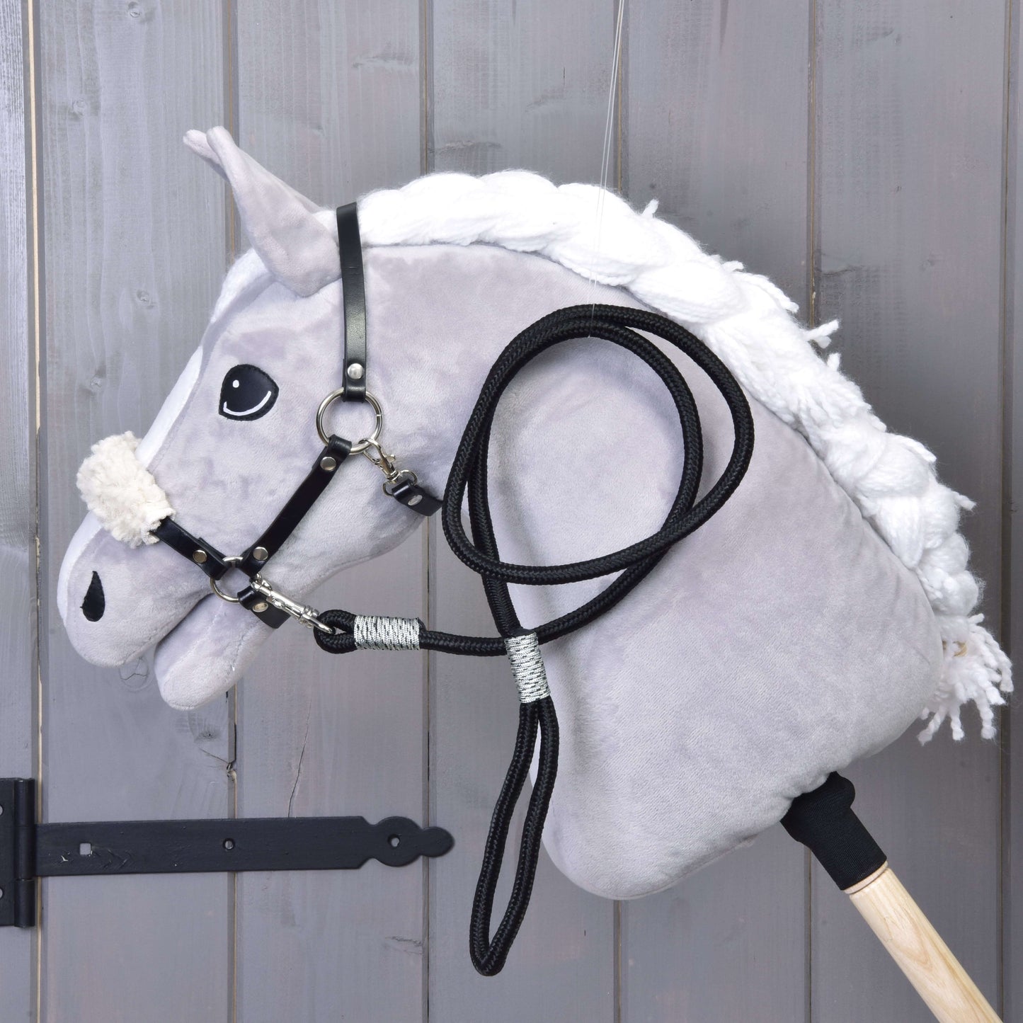 Hobby Horse LarDen Set Basic zobrazuje plyšového Hobby Horse s bielou hrivou, čiernou uzdečkou s opratami a čiernou ohlávkou na drevenej tyči, opretého o sivé drevené panely.