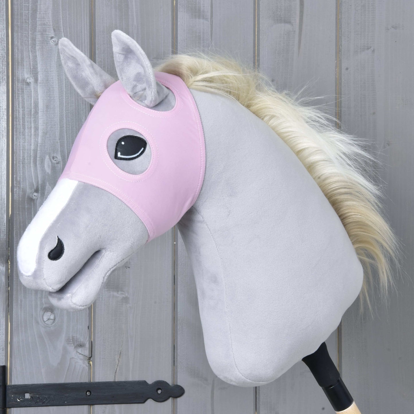 Plyšový Hobby Horse od Hobby Horse LarDen má nasadenú Masku bez uší Pink – prateľnú ružovú masku – na svetlosivom tele, s bielym mulcom a krémovou hrivou. Kôň je nasadený na drevenej tyči na pozadí sivej drevenej steny.