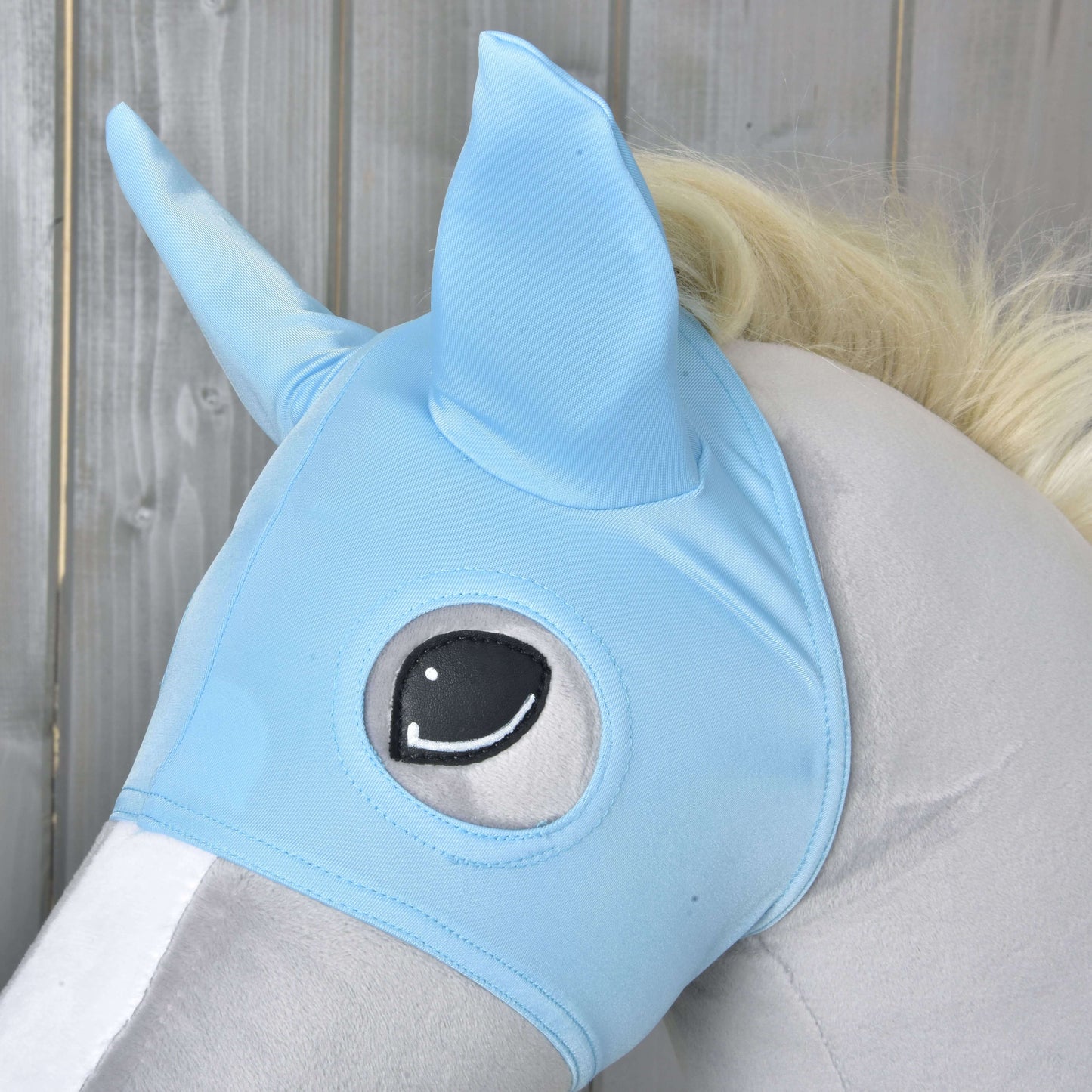 Detail plyšovej hračky koňa s Maska Sky od Hobby Horse LarDen, so špicatými ušami a ilustrovaným okom, na drevenom pozadí.