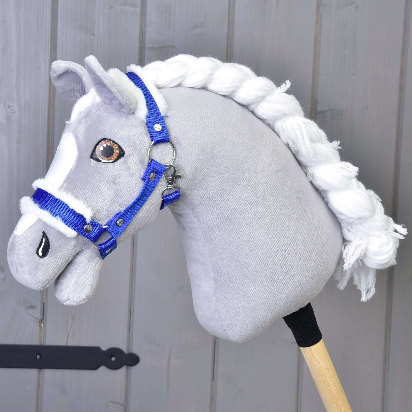 Sivý látkový Hobby Horse s bielou priadzovou hrivou a ohlávkou Hobby Horse LarDen Ohlávka modrá s barančekom stojí pred svetlosivou drevenou stenou.
