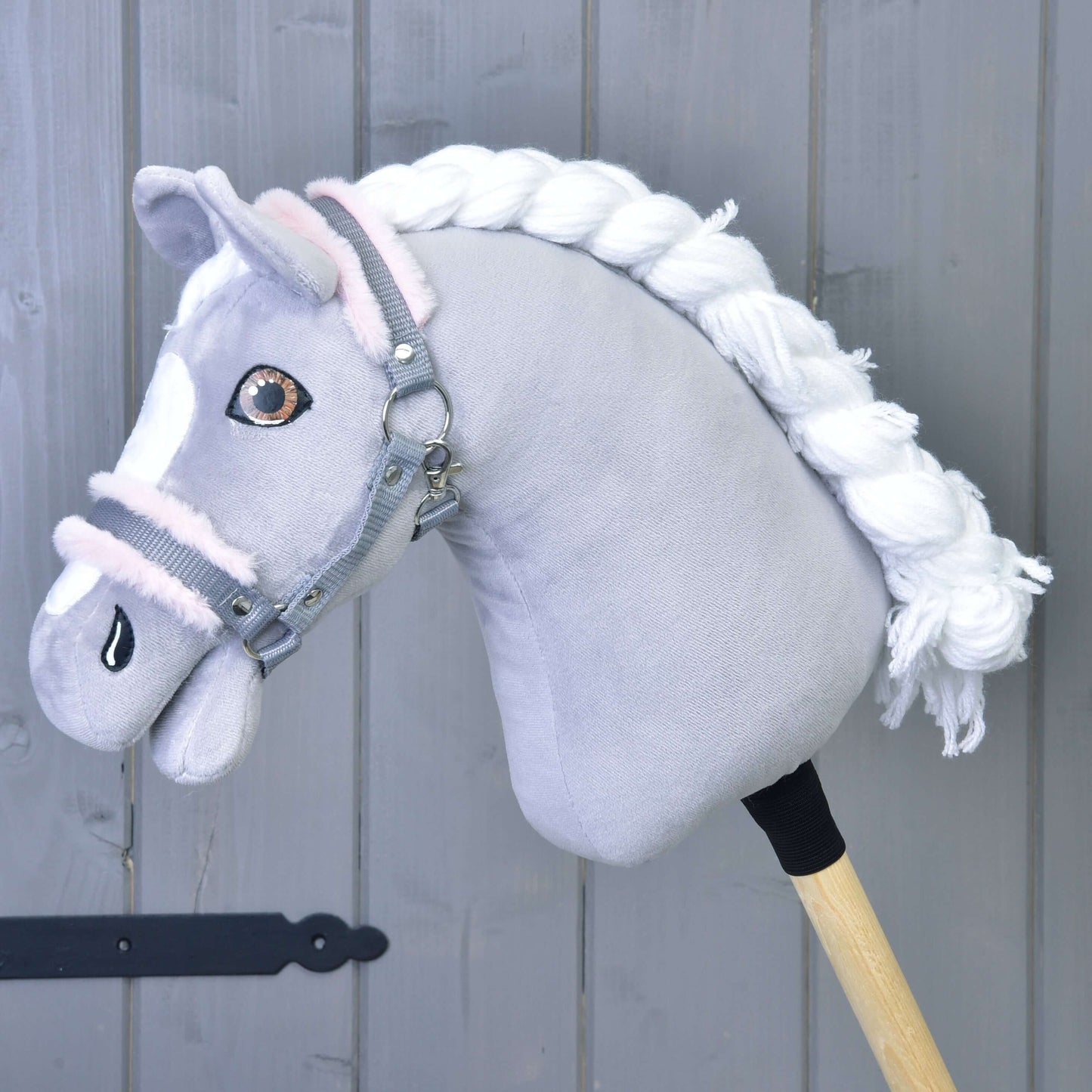 Hobby Horse LarDen "Ohlávka sivá s pink barančekom" zobrazuje sivého plyšového koňa s bielou hrivou a sivou ohlávkou, pripevneného k drevenej tyči, na pozadí sivých drevených panelov.
