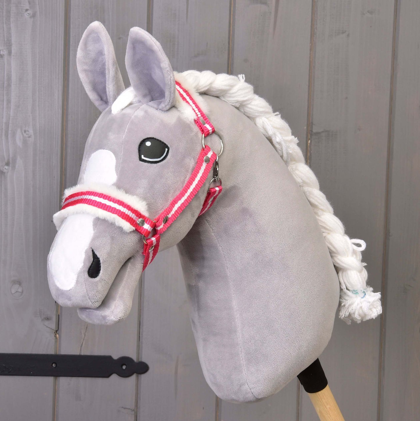 Hobby Horse LarDen "Ohlávka s červeným prúžkom a barančekom" je sivý plyšový Hobby Horse s bielou hrivou a ohlávkou s červeným prúžkom na drevenej tyči, zobrazený pred sivou drevenou stenou.