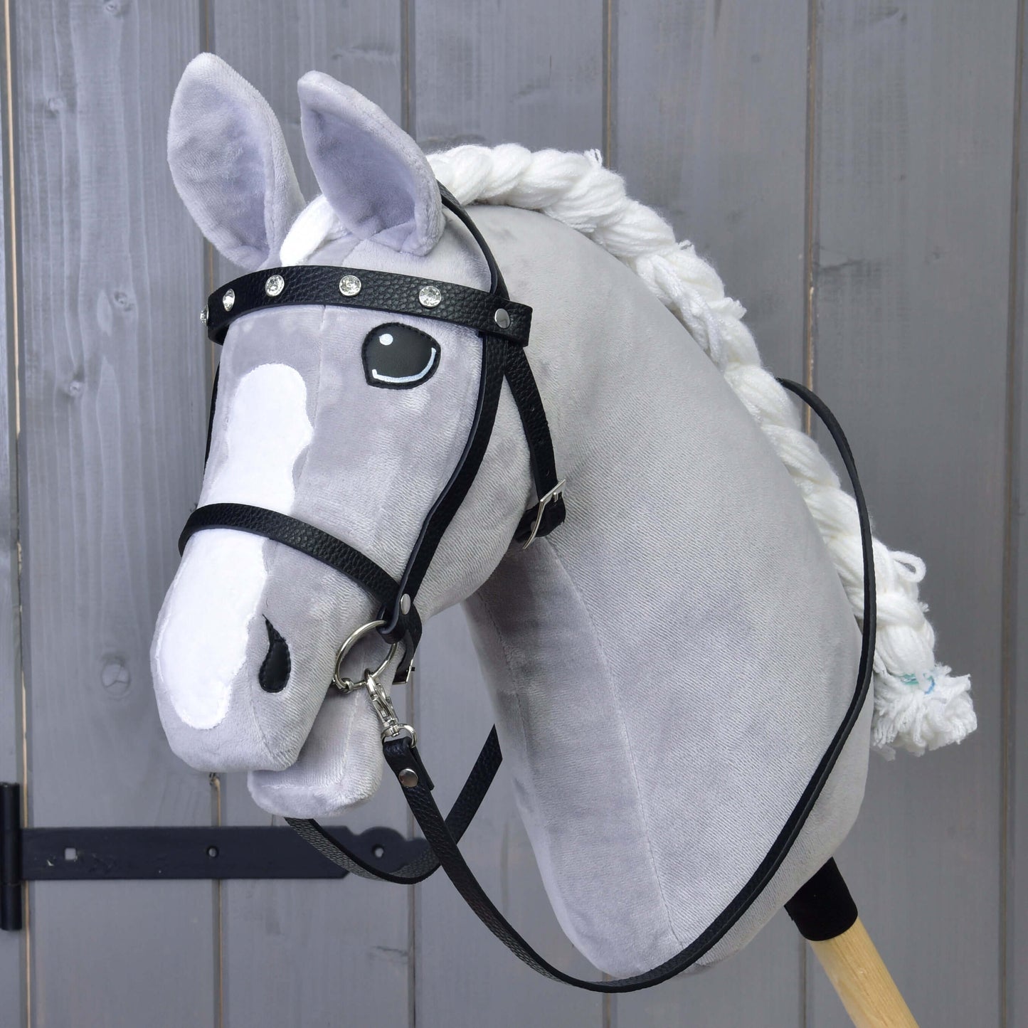 Šedý plyšový Hobby Horse od Hobby Horse LarDen s Kompletní uzdečkou s kamínky z eko kůže, bílou hřívou, odnímatelnými otěžemi a dřevěnou tyčí, před šedou dřevěnou stěnou.